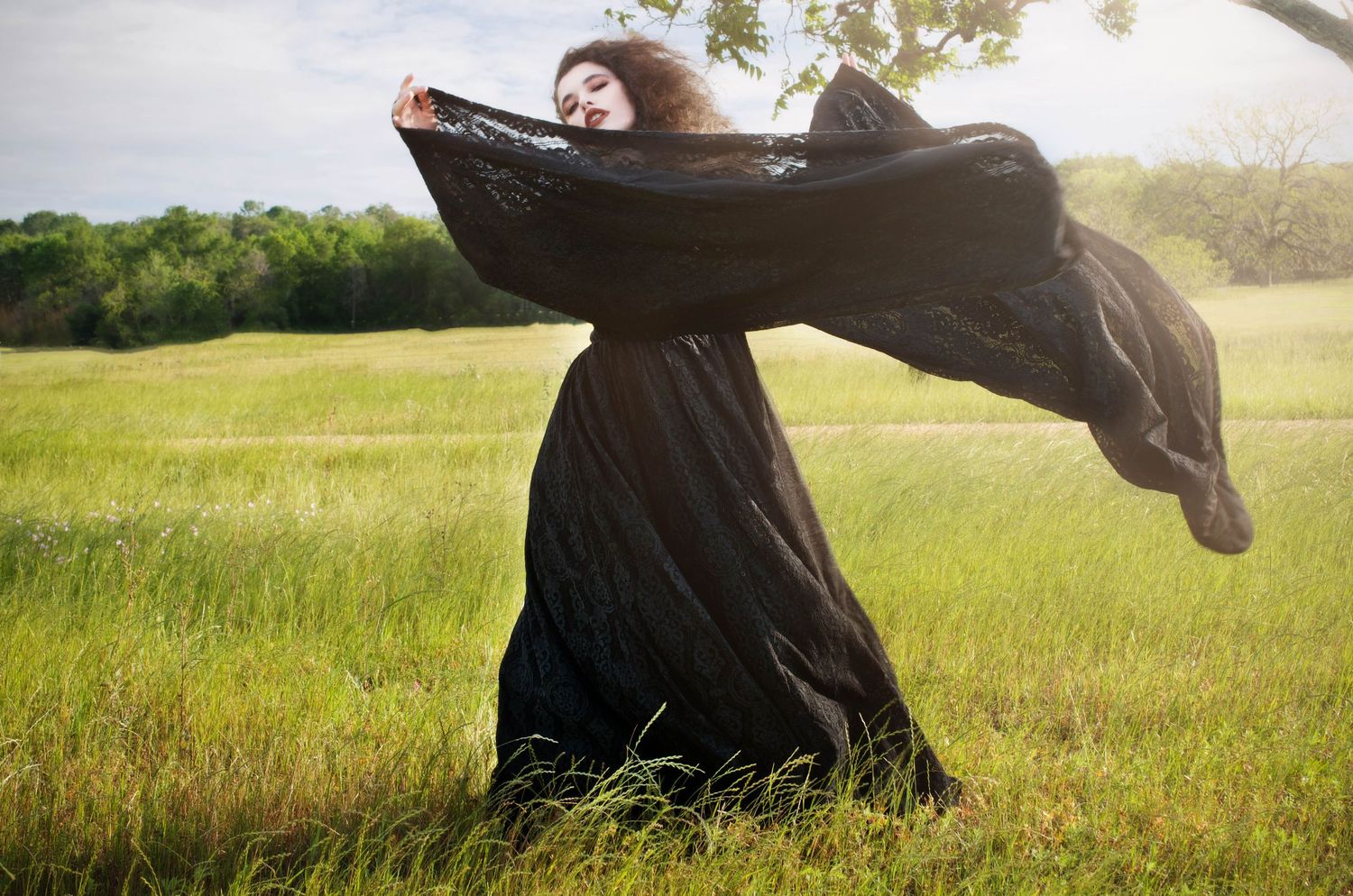 Raven haired beauty stands in a field in Texas wearing a black dress, flowing sleeves floating on the wind.