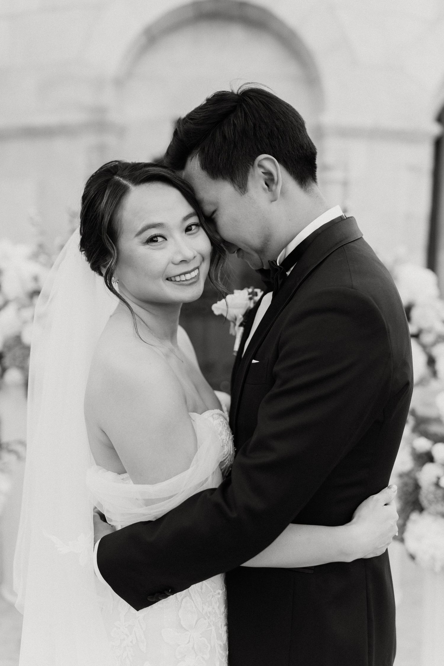Black and white photo of a smiling bride in a veil embracing a groom in a tuxedo at a wedding ceremony.