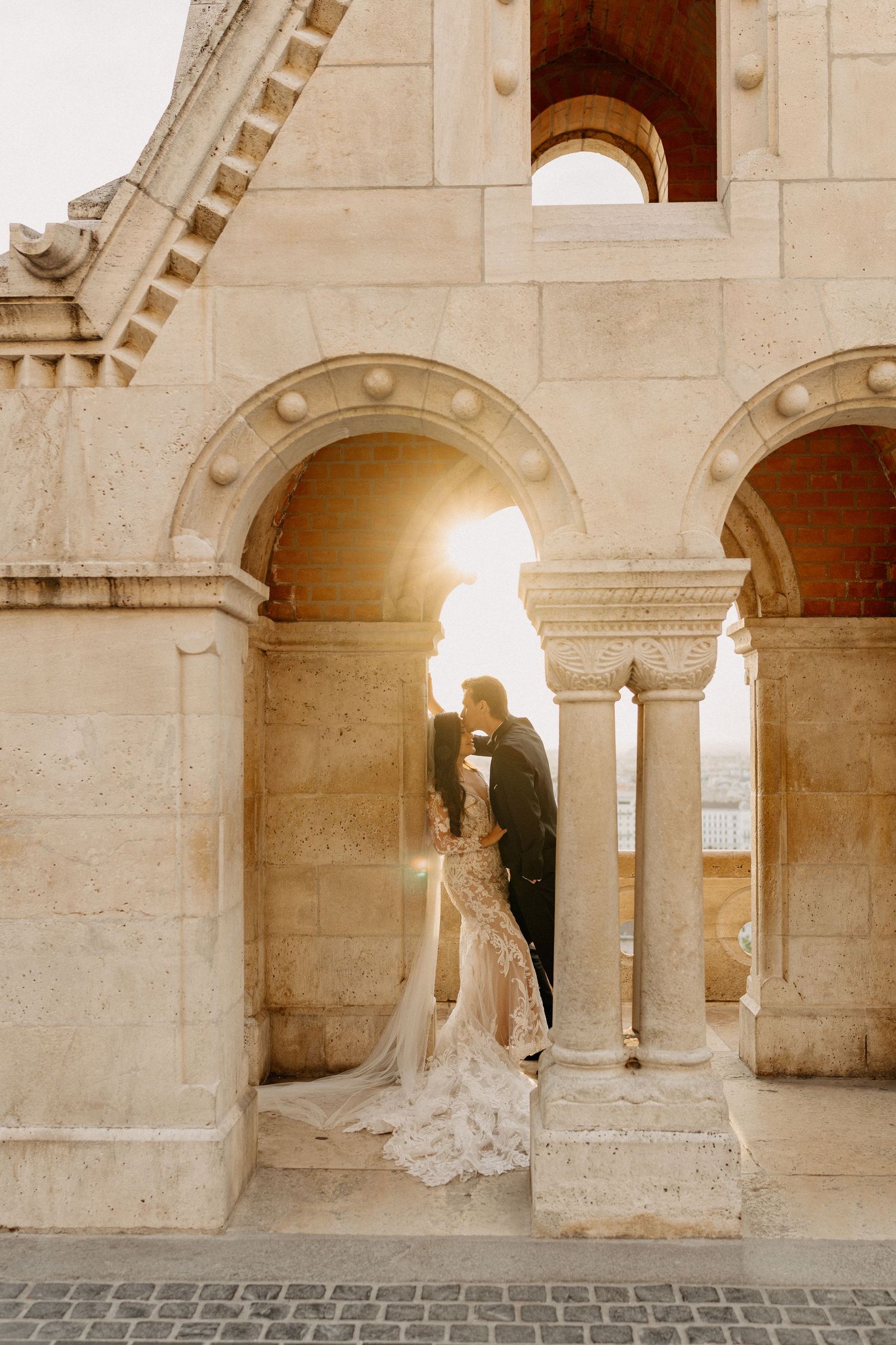 Bride and groom kissing at sunset beneath stone arches at Fisherman's Bastion, Budapest, in elegant wedding attire.