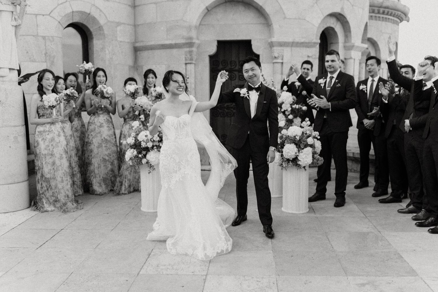 Black and white photo of a joyful couple exiting their wedding ceremony surrounded by bridesmaids and groomsmen.