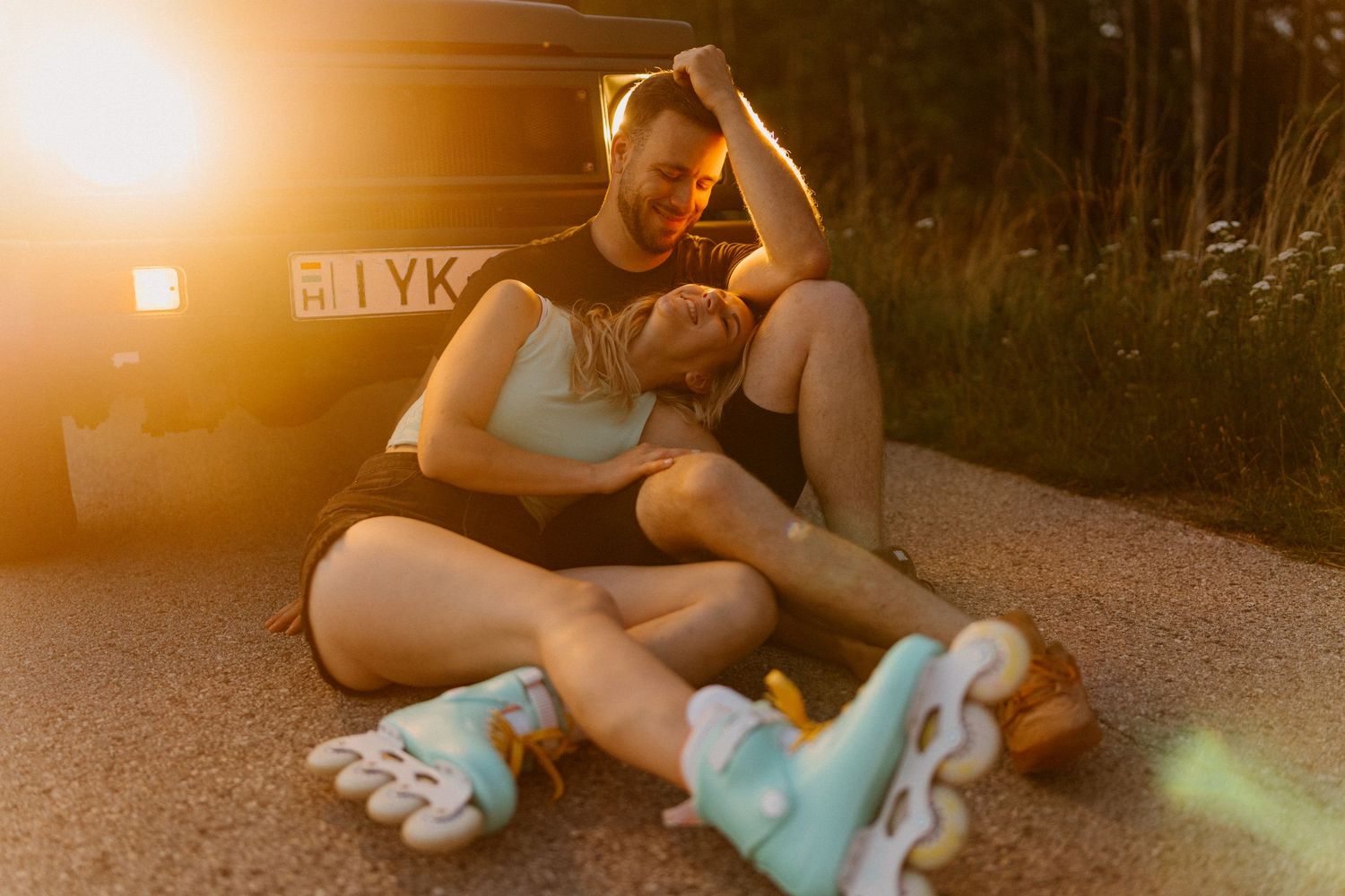 Couple sitting together on ground, wearing inline roller skates, leaning against a car in warm golden light.