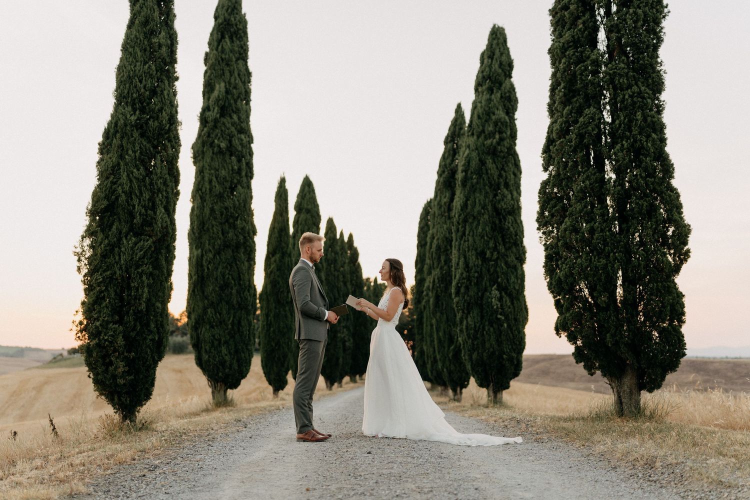 Bride and groom hold hands on gravel path lined with tall cypress trees at Tuscan sunset.