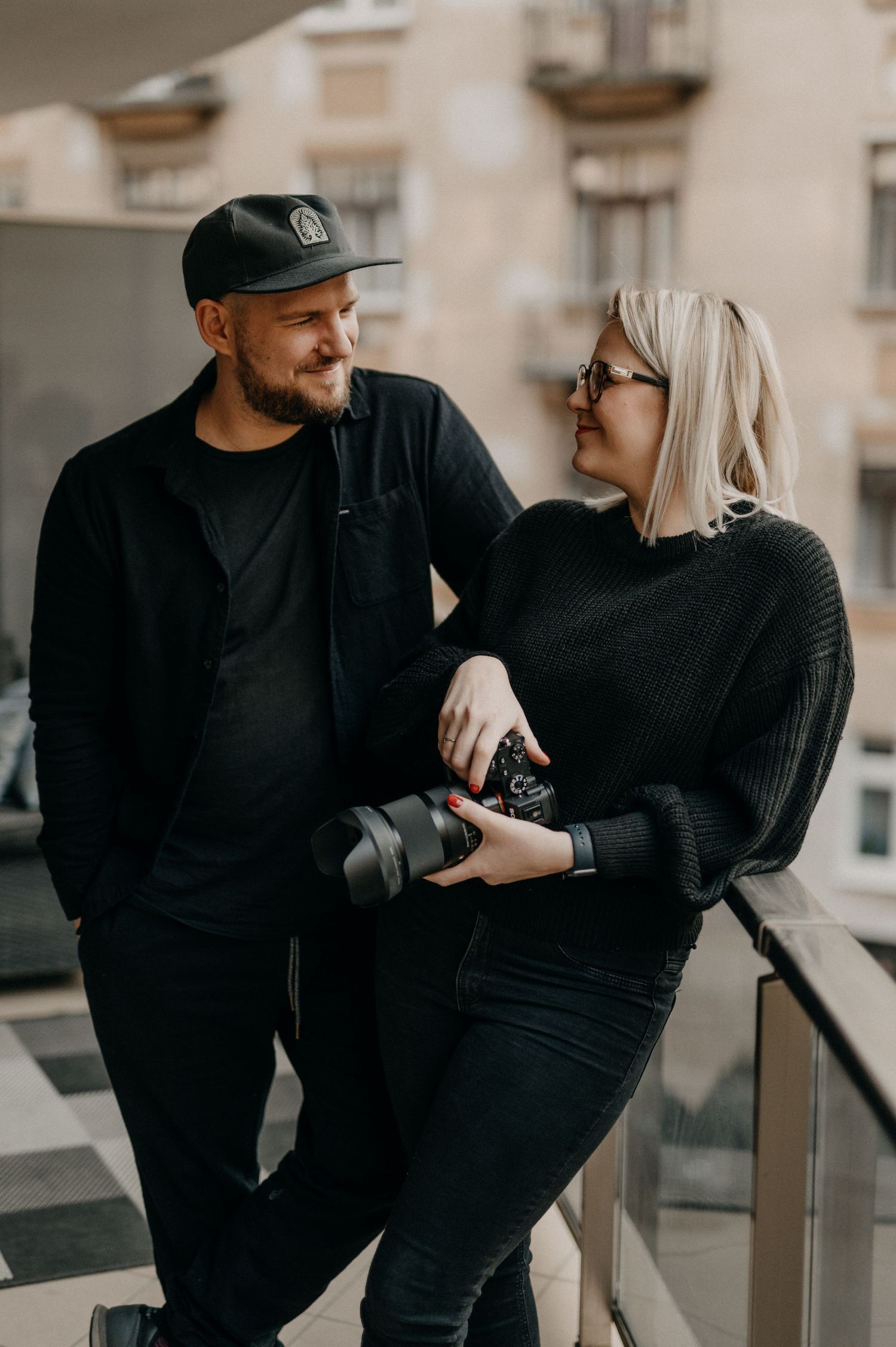Male and female photographer duo smiling at each other on a balcony, woman holding a camera.