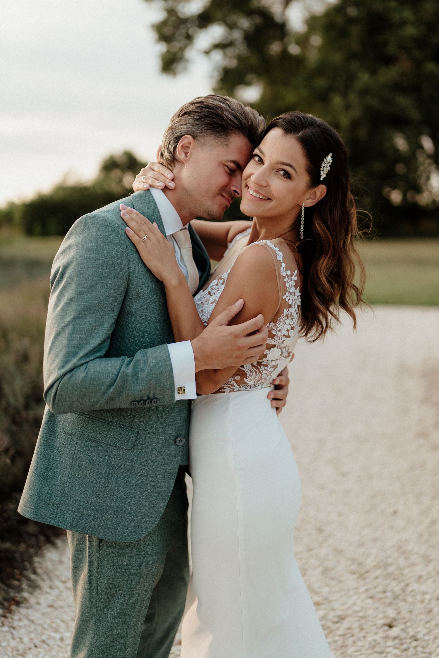 A bride in a white lace gown smiles as her groom in a teal suit embraces her on a gravel path.