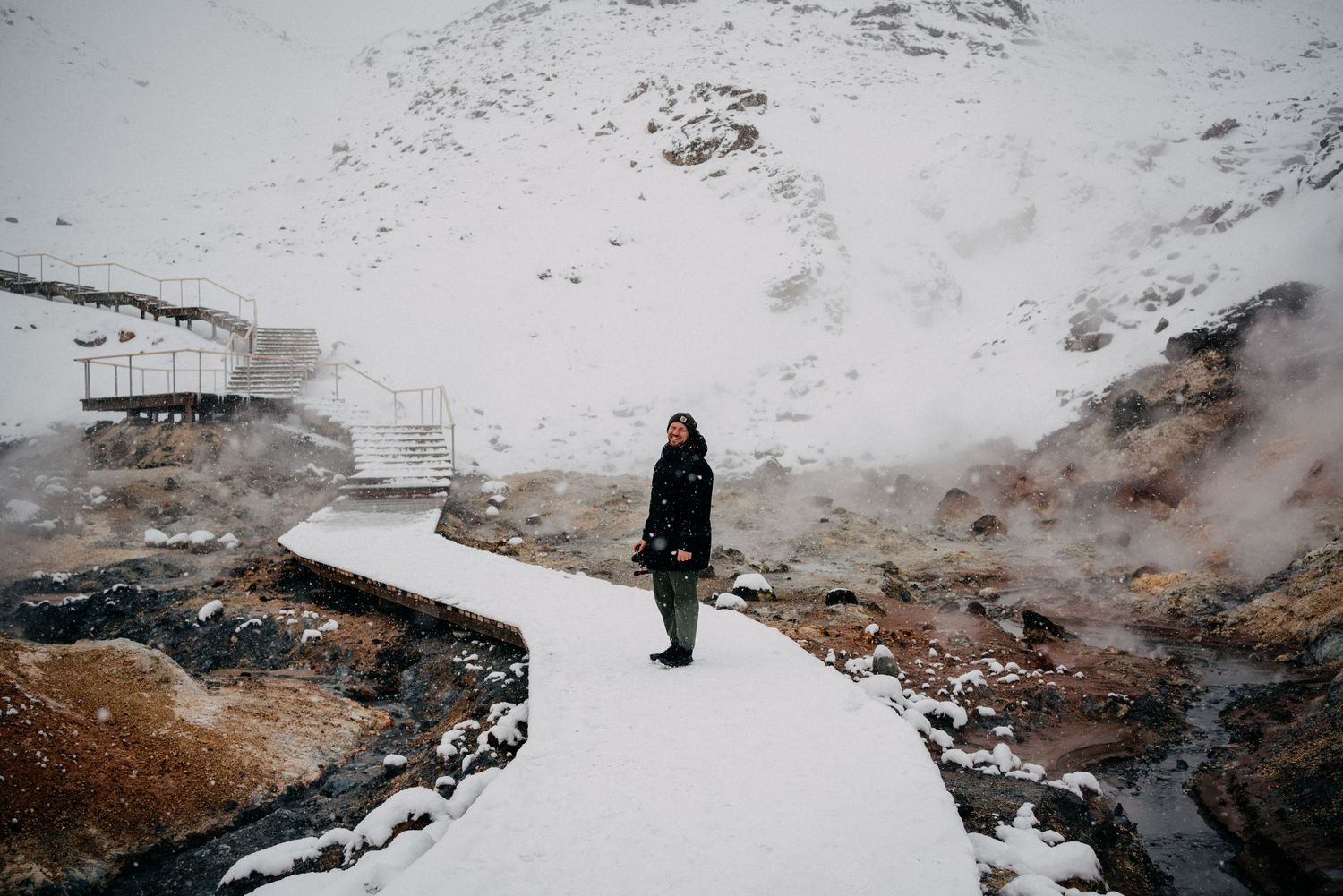 Woman stands on snowy geothermal hot spring boardwalk amid steaming volcanic landscape in Iceland.