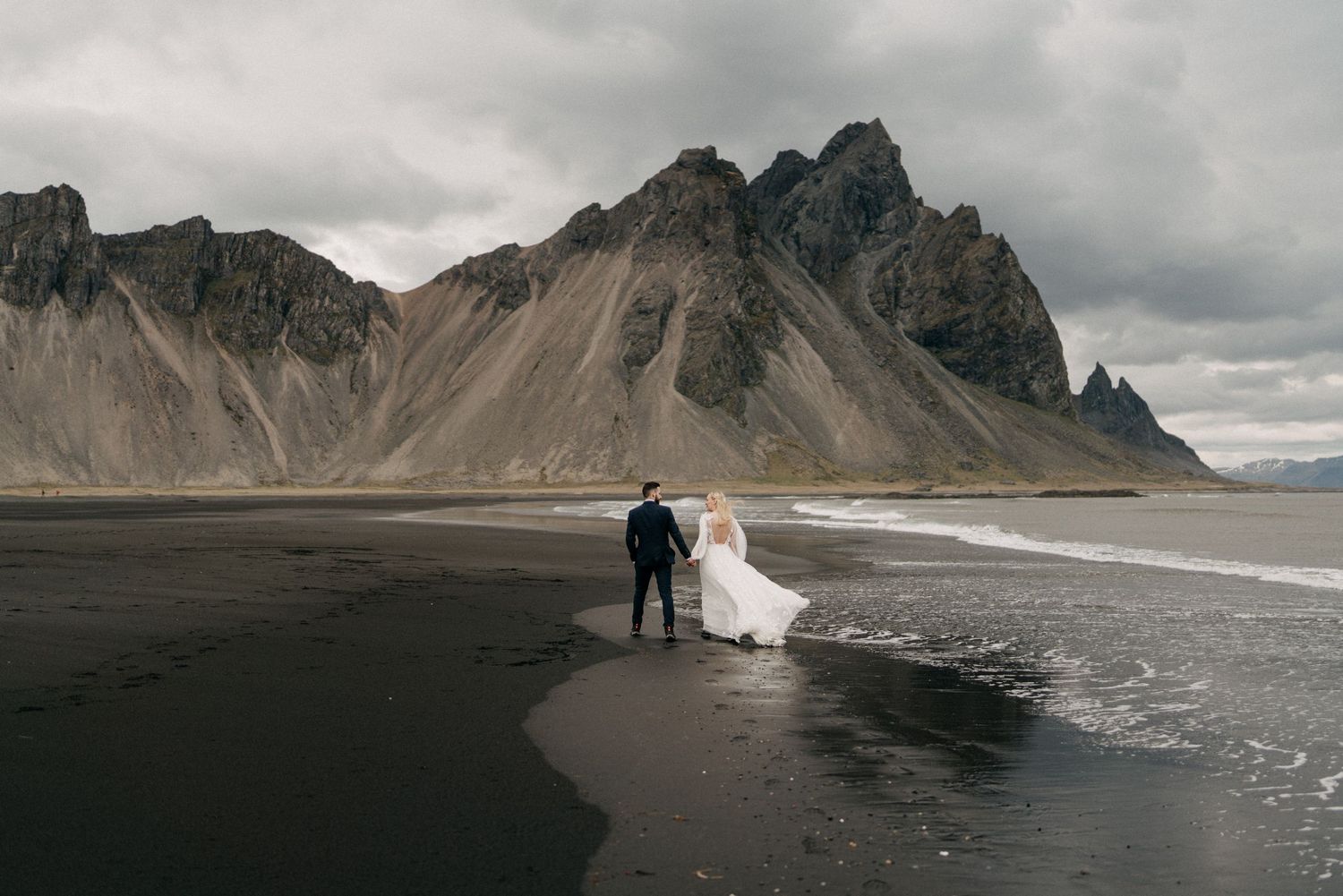 Bride and groom hold hands on dramatic black sand beach with rugged Icelandic mountains in background.