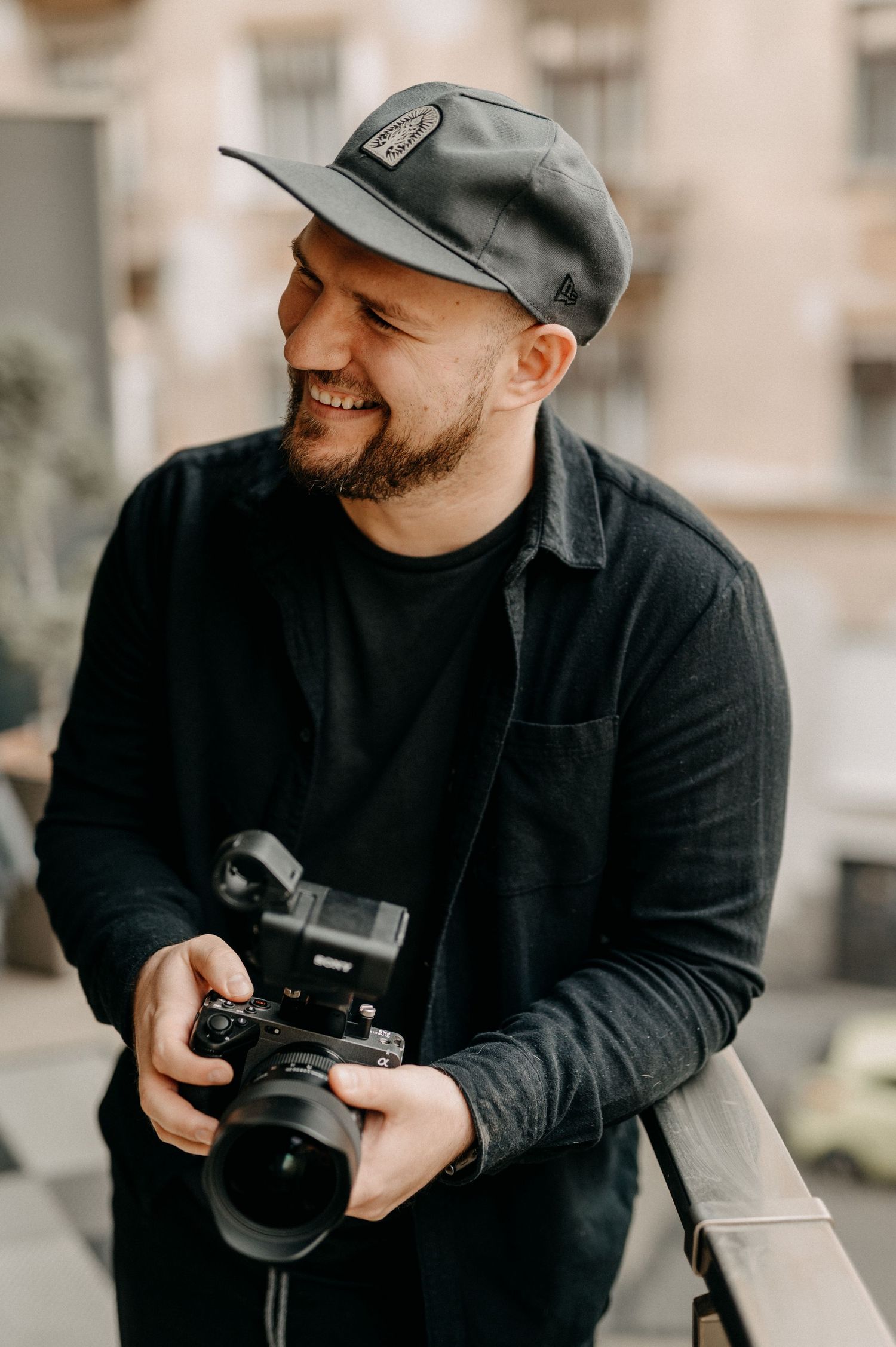 Smiling male photographer in black outfit and cap holding a mirrorless camera on a balcony.