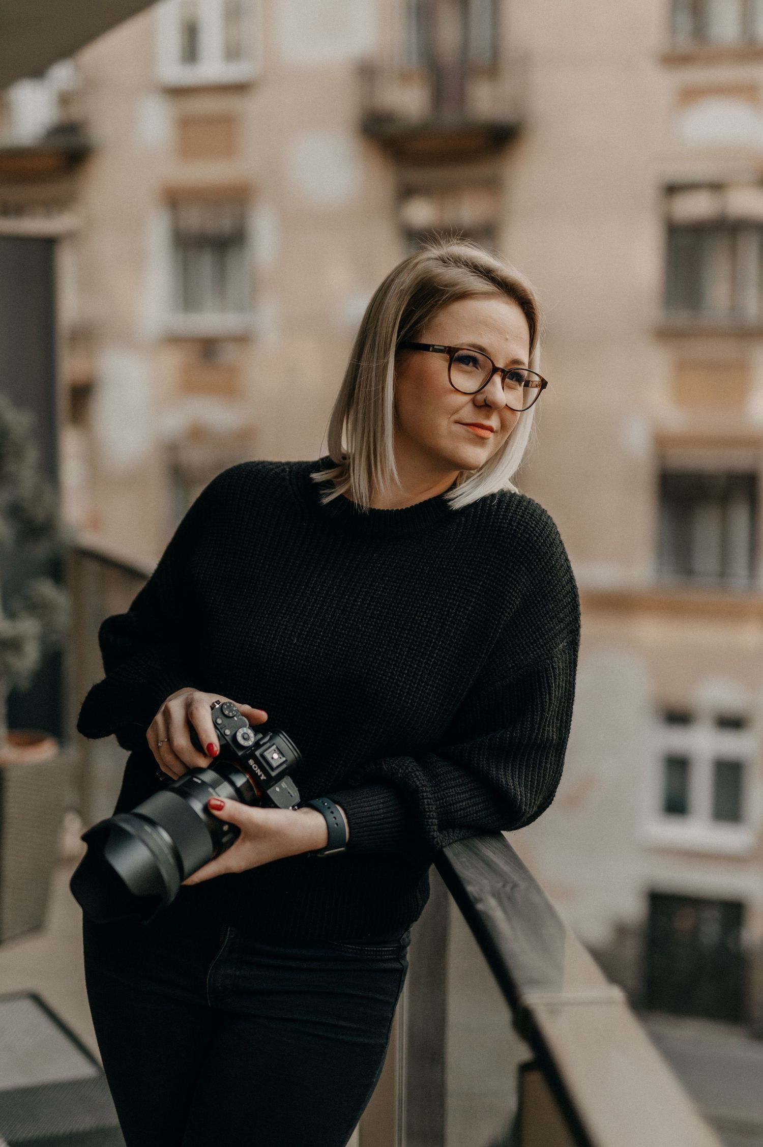 Blonde female photographer in black sweater holding a camera while gazing from a balcony.