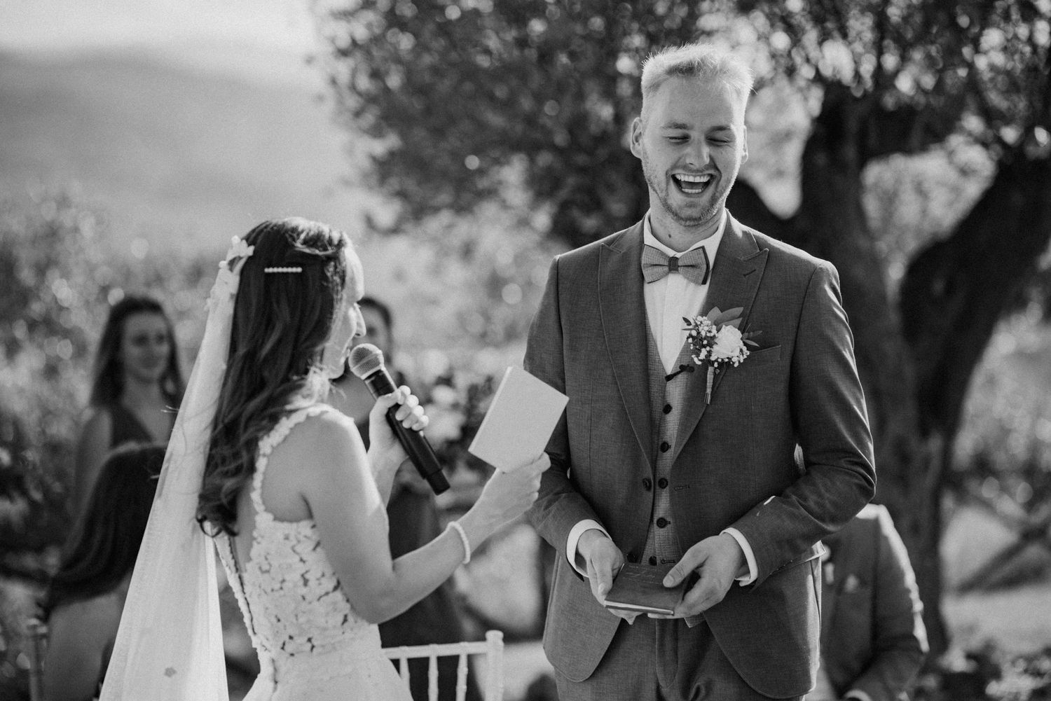 Groom laughing joyfully as bride reads wedding vows during outdoor ceremony, black and white photo.