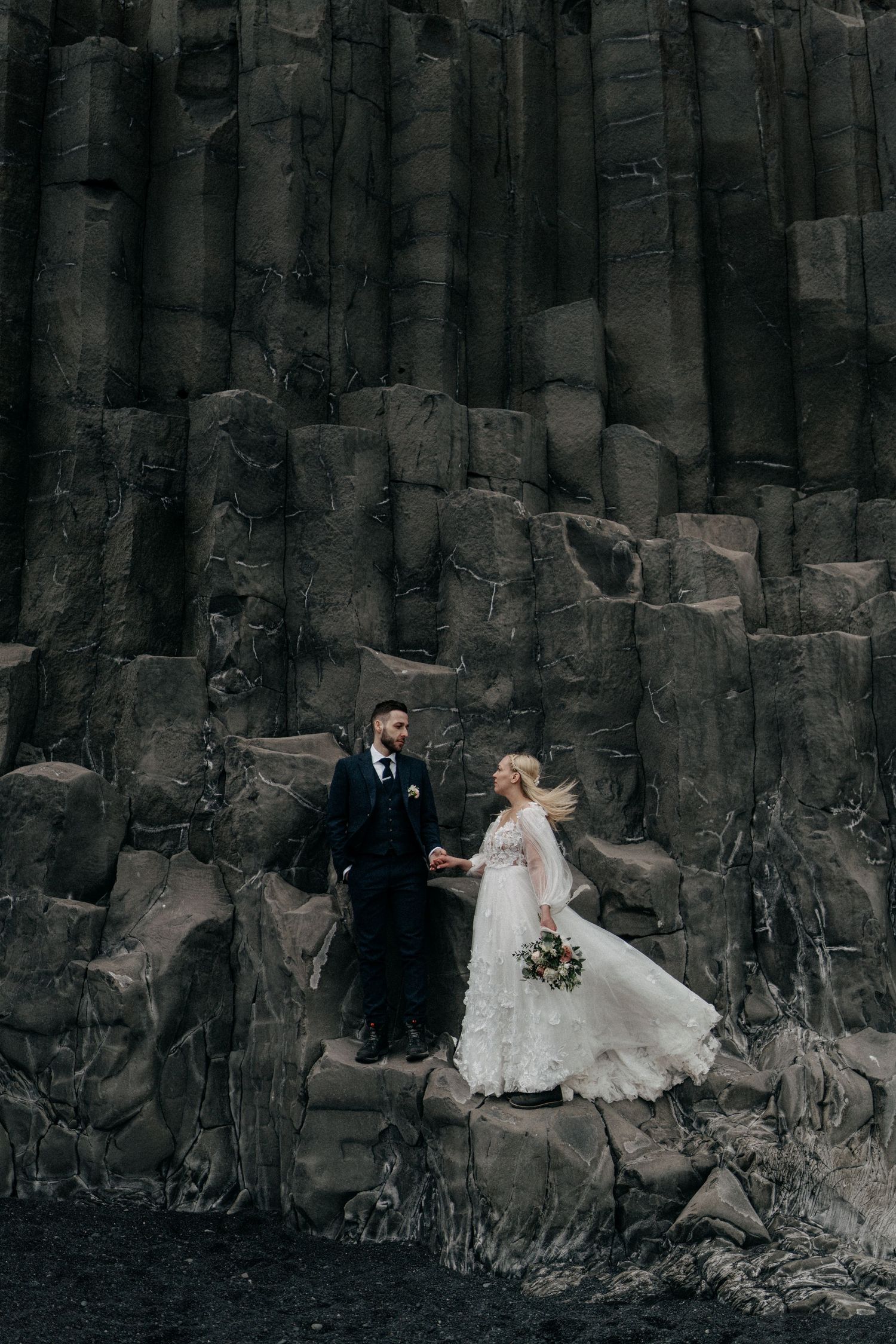 Bride and groom holding hands beside dramatic black basalt columns at Iceland black sand beach.