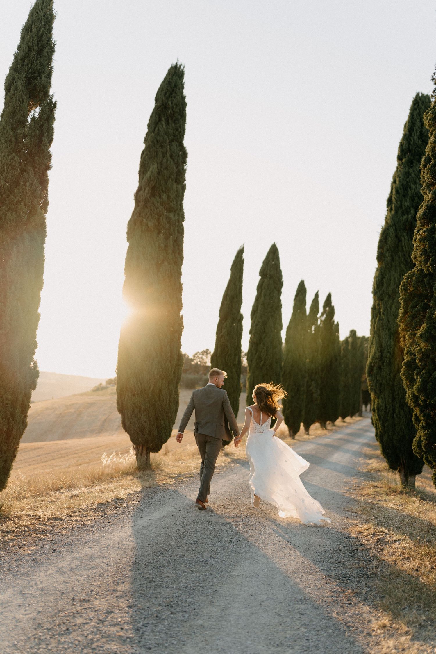Bride and groom walk hand-in-hand down cypress-lined Tuscan road at golden sunset.