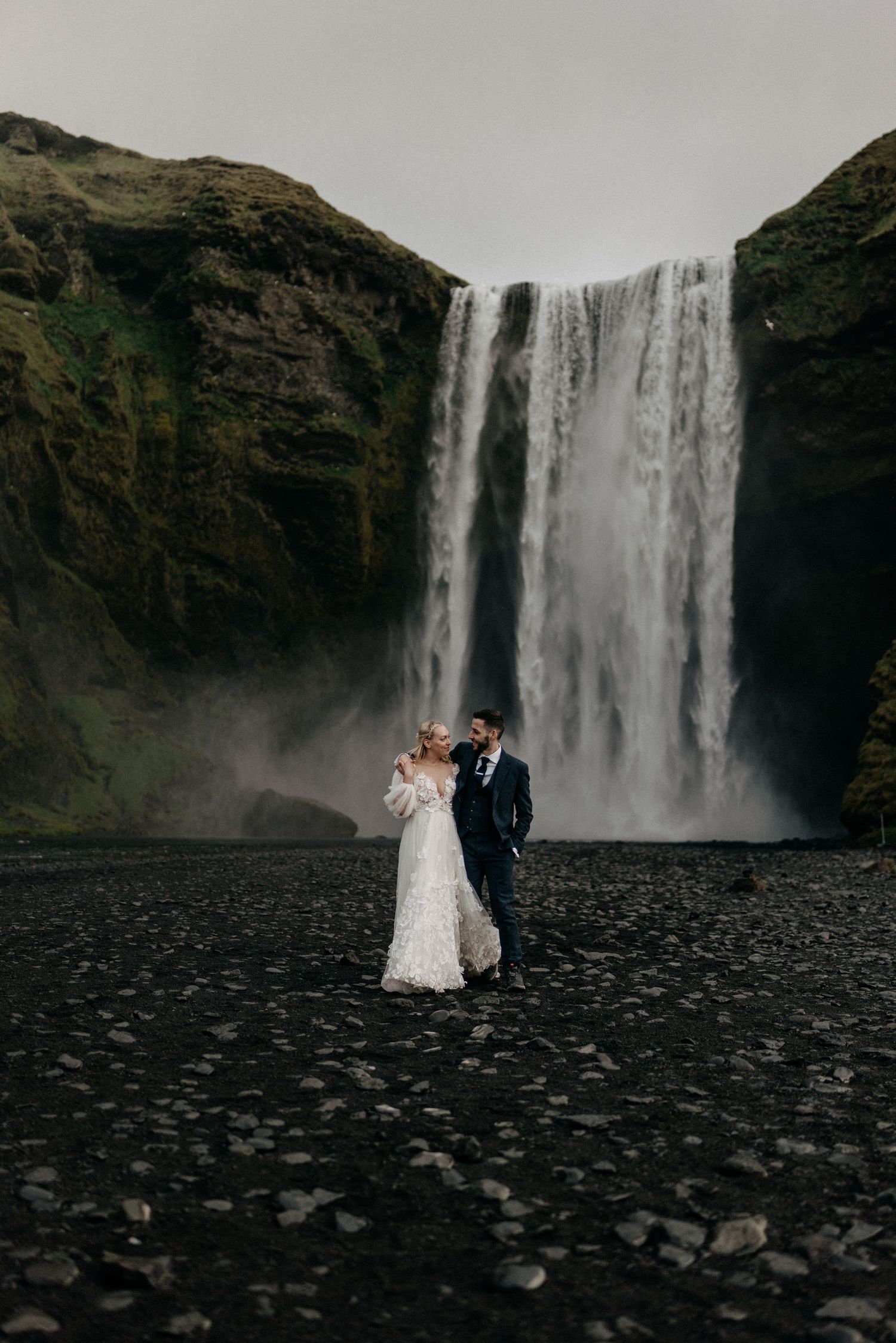 Wedding couple embraces before majestic Skogafoss waterfall in Iceland on their elopement day.
