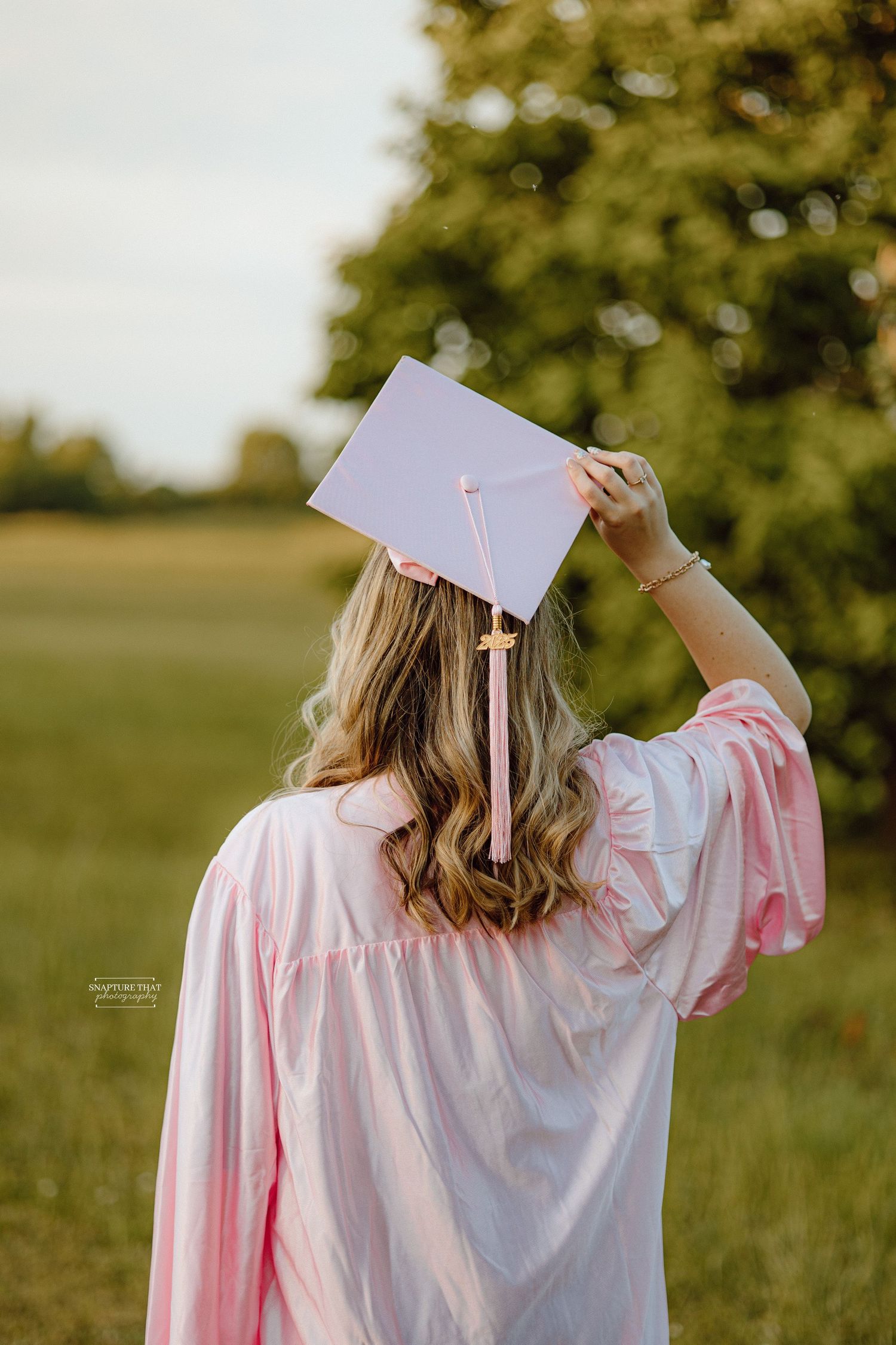Graduate in pink gown holds up diploma against green outdoor background.
