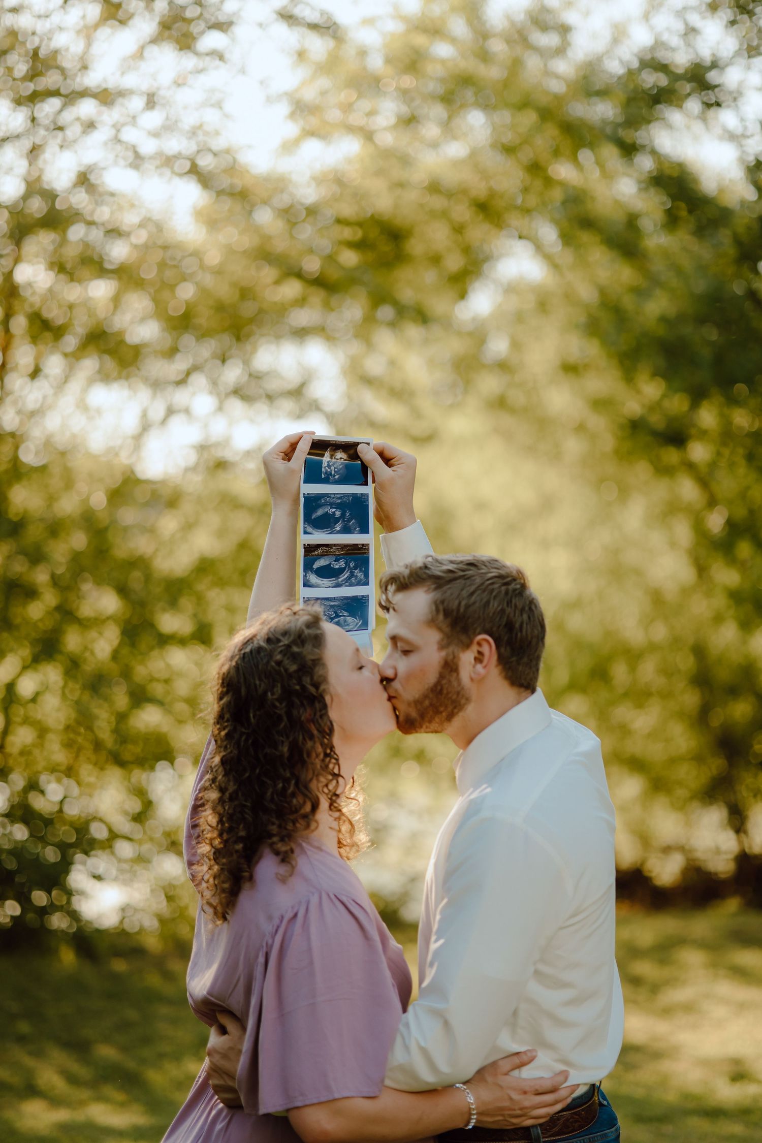 A couple shares a kiss while holding ultrasound photos in golden sunlight.