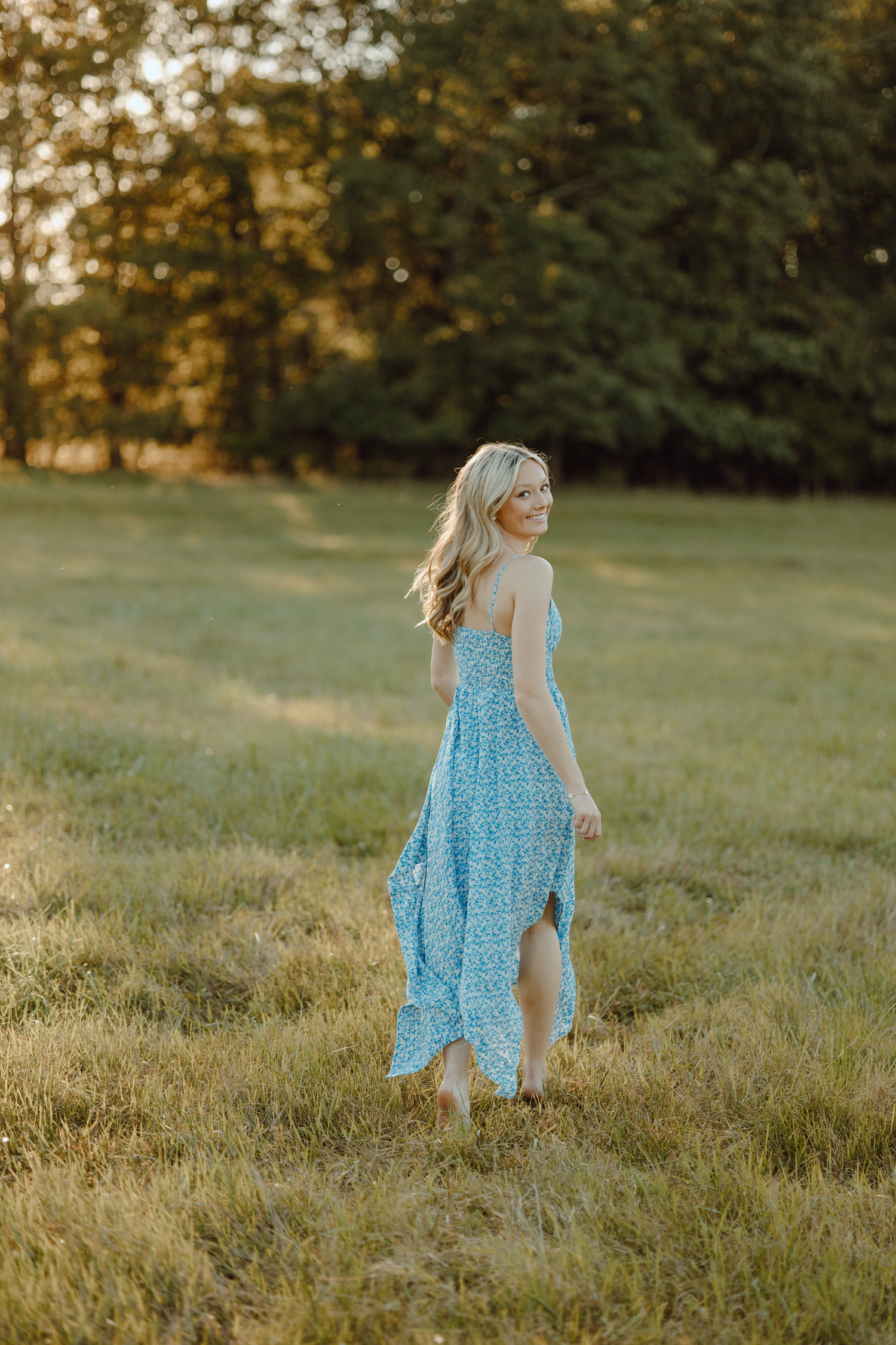 Someone wearing a blue floral dress walking through a sunlit meadow at sunset.
