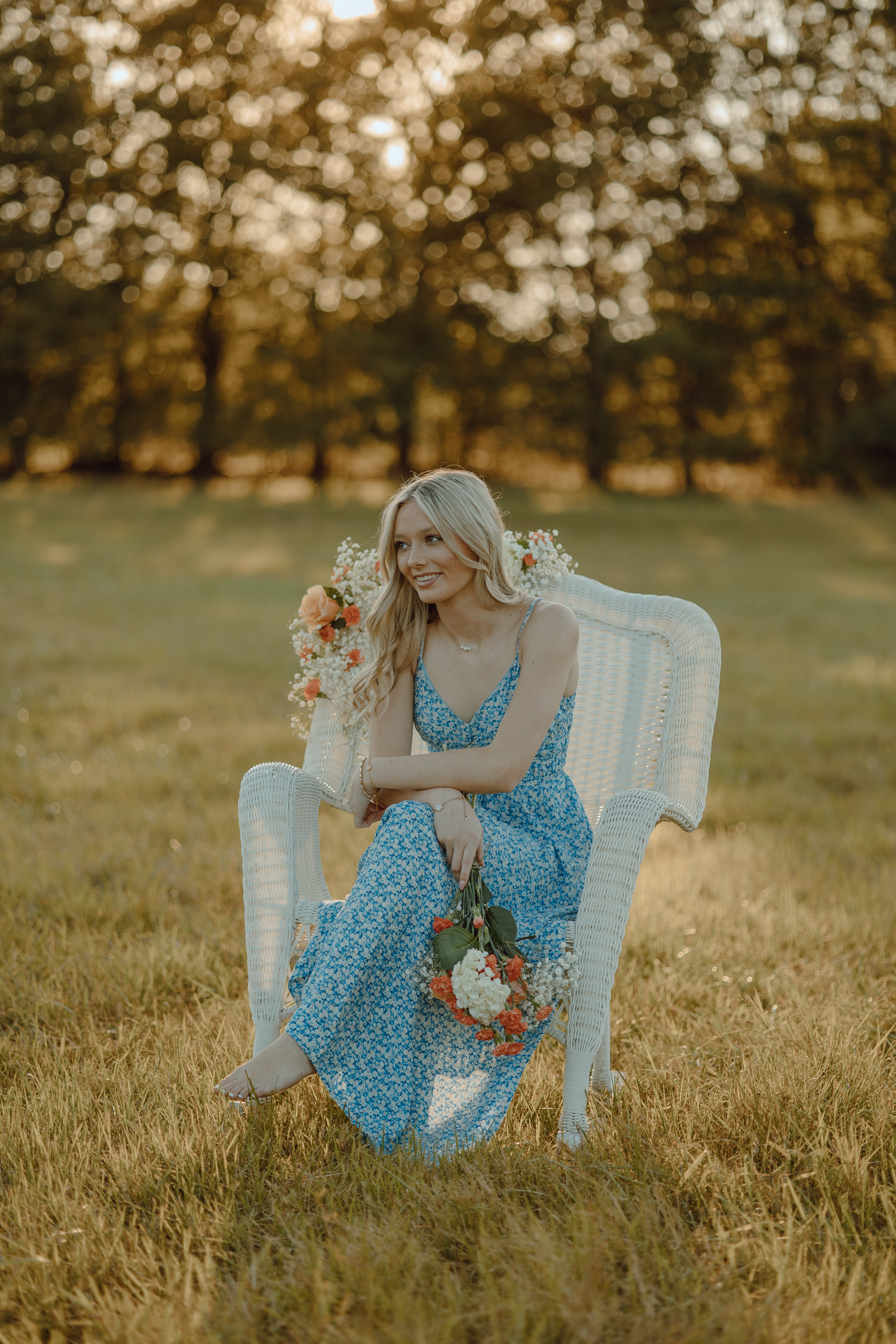 Woman in blue floral dress poses on white vintage chair in sunlit field.