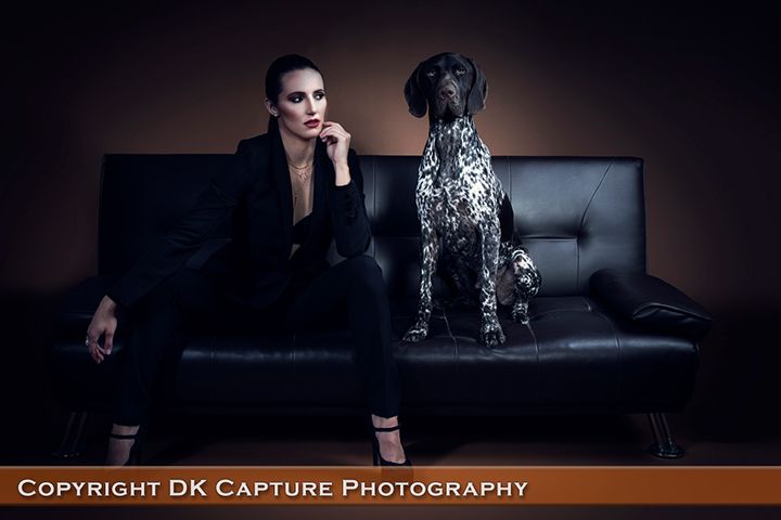 A stylish portrait featuring a person in dark attire seated on a black leather couch with a German Shorthaired Pointer dog.