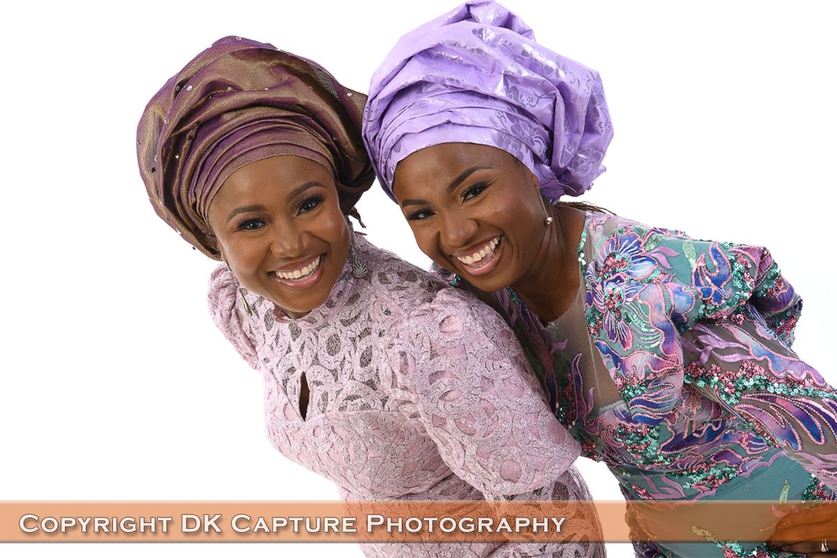 Two people wearing colorful traditional African head wraps and clothing smile while leaning together.