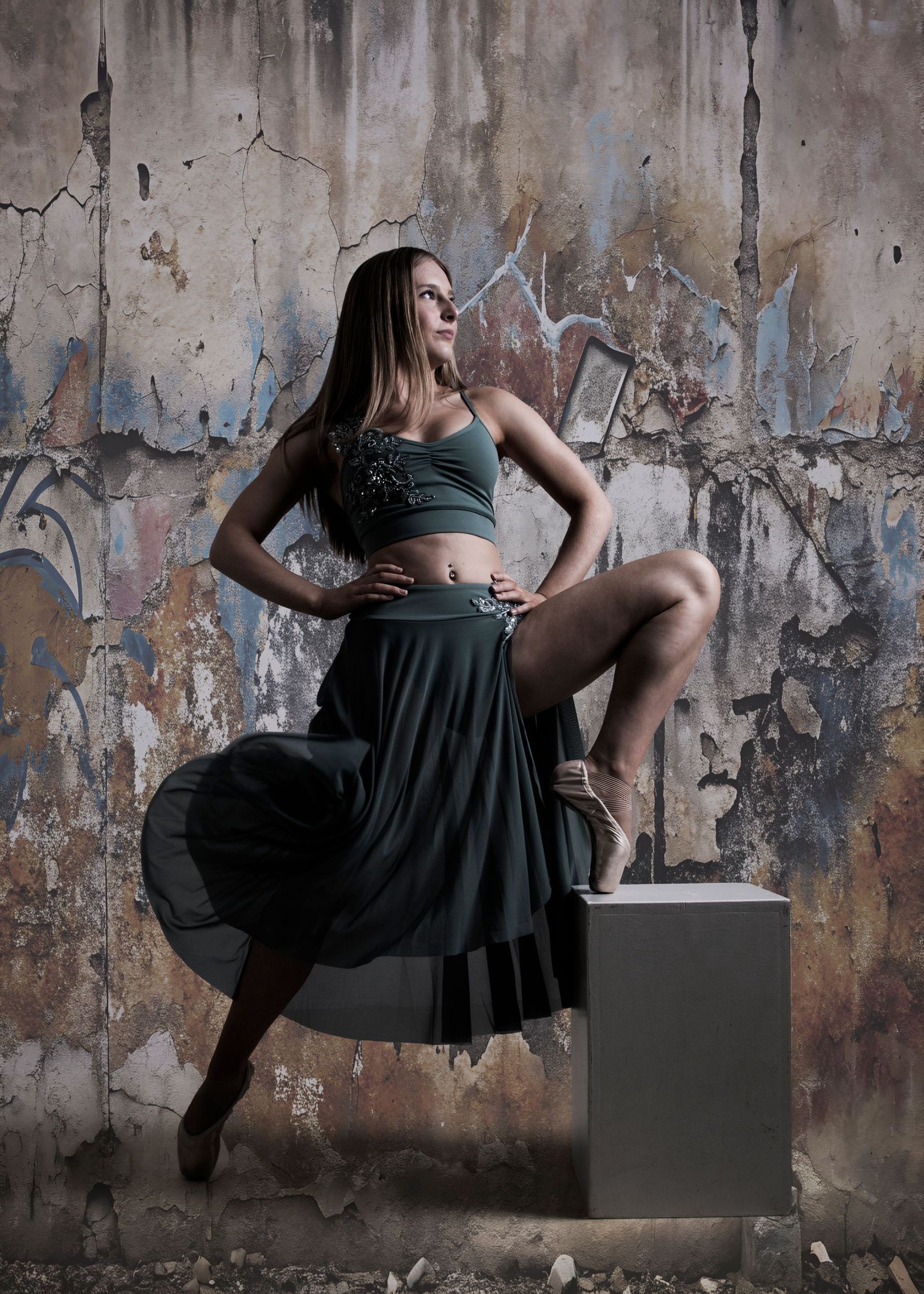 A dancer in a teal crop top and skirt poses gracefully on a cube against a graffiti-covered wall.