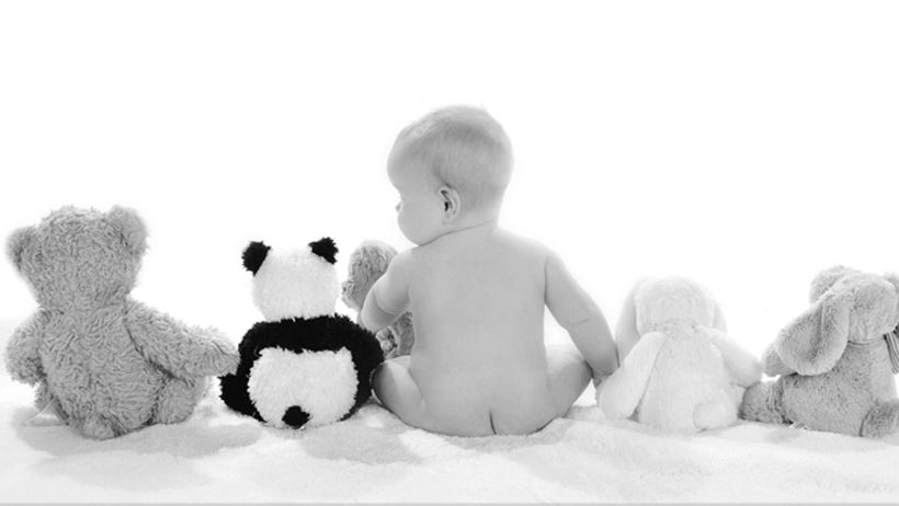 Black and white photo of a baby sitting with stuffed animal toys including a panda and teddy bears.