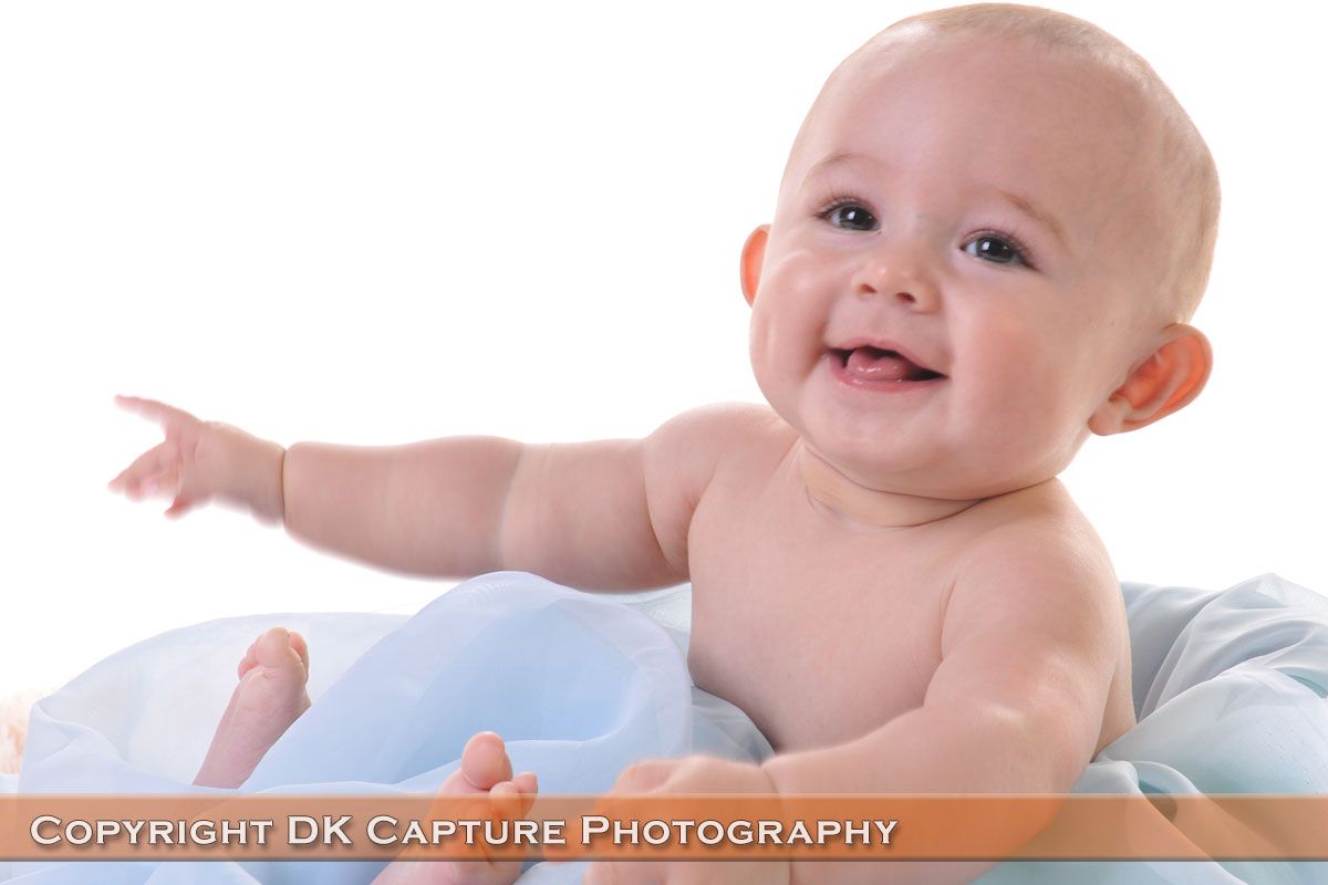 A baby with blonde hair wearing a light blue shirt lies on a white blanket and smiles brightly at the camera.