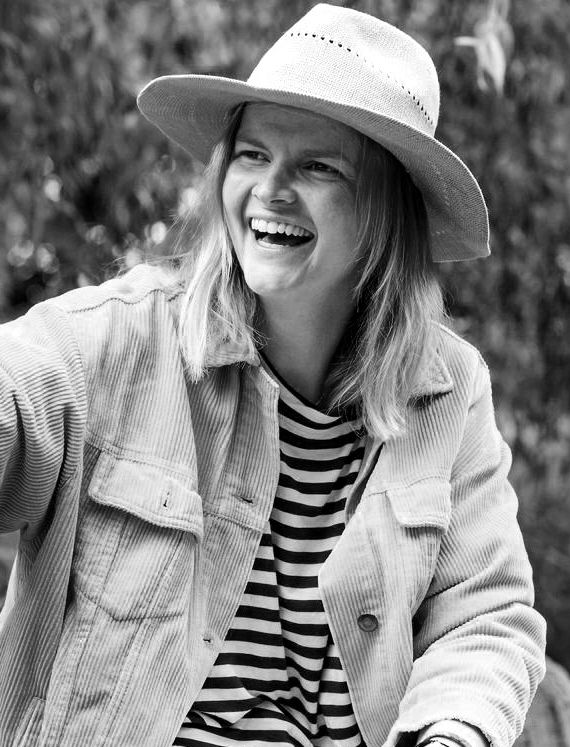 A laughing woman wearing a straw hat, striped shirt, and corduroy jacket outdoors in a black and white photo.