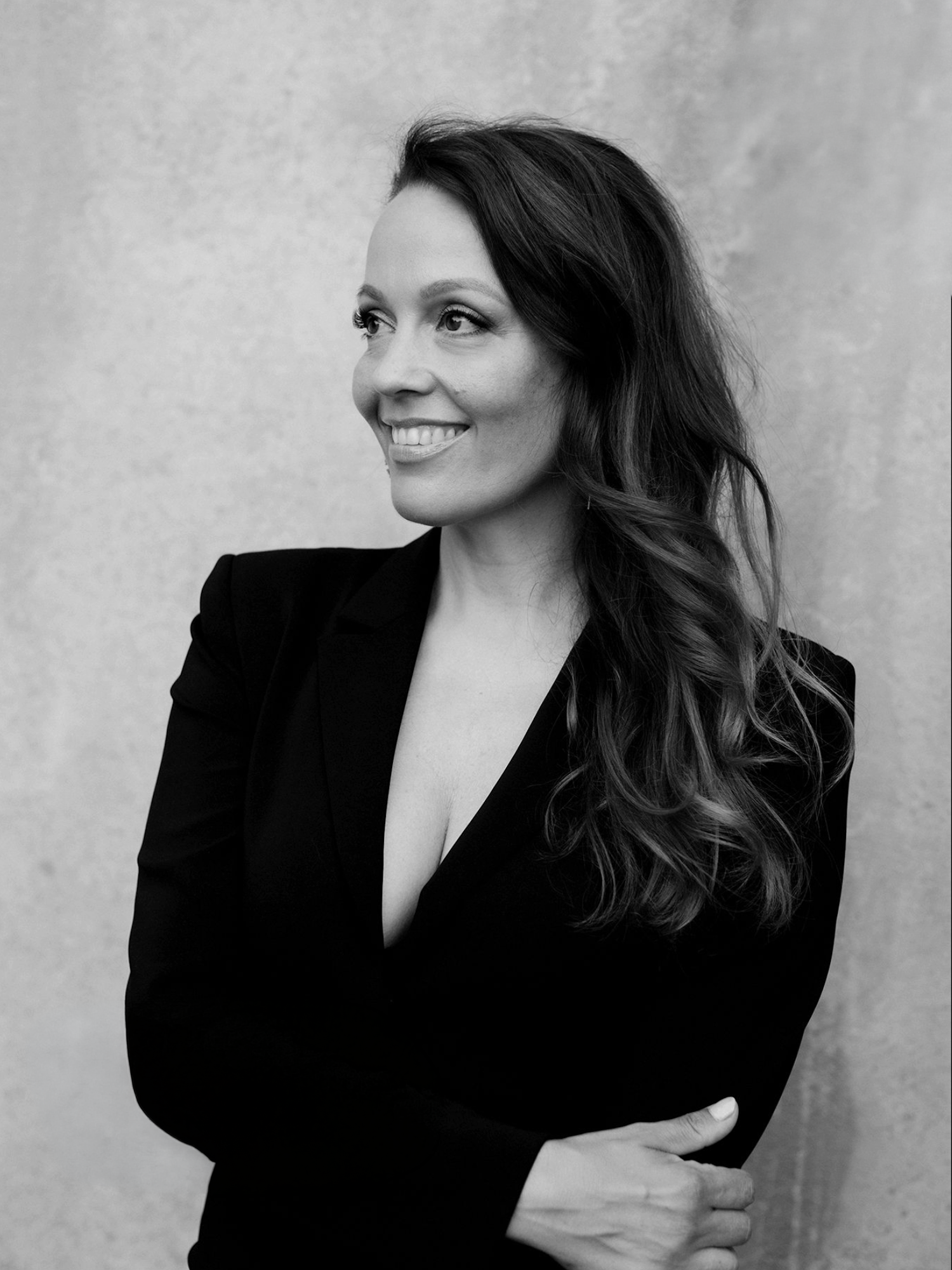 Black and white portrait of a smiling woman with long hair in a black blazer, arms crossed.
