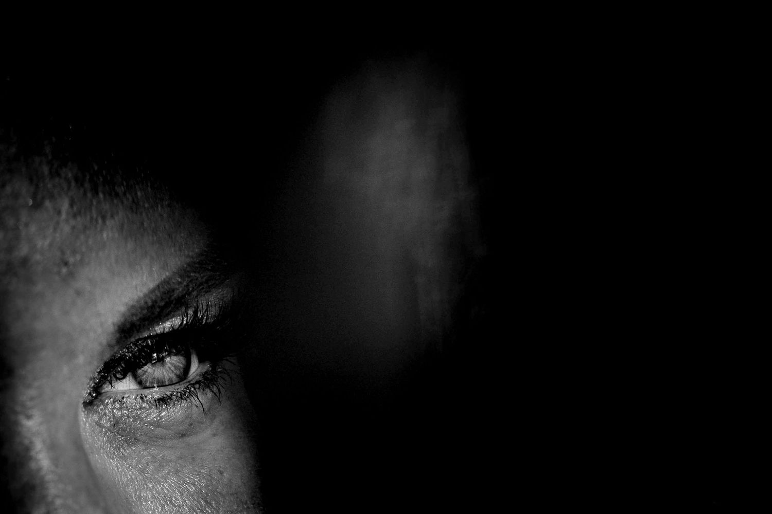 Dramatic black and white close-up photograph of a person's eye emerging from darkness with intense contrast.