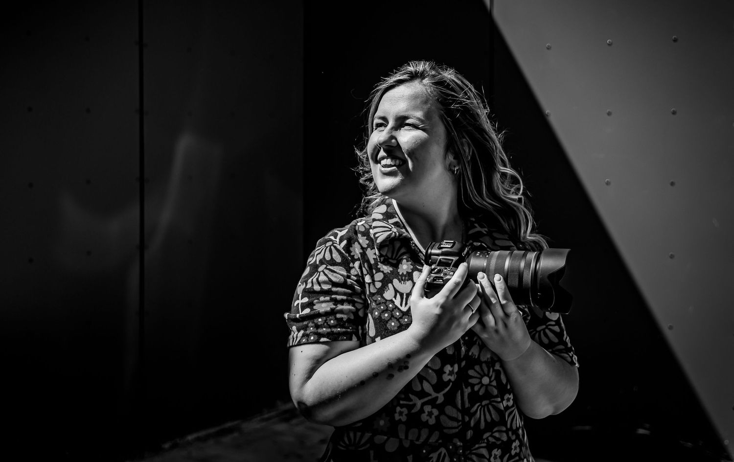 Black and white photo of a smiling woman holding a camera outdoors, wearing a floral shirt.
