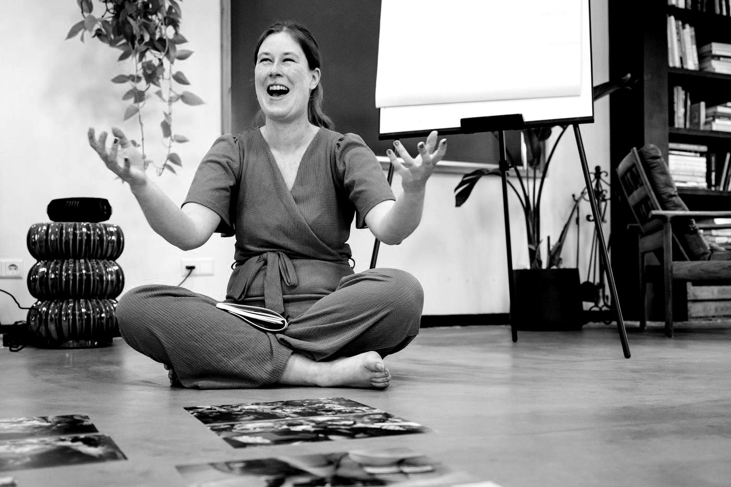 Woman sitting cross-legged on floor laughing with hands raised in a yoga studio, black and white photo.