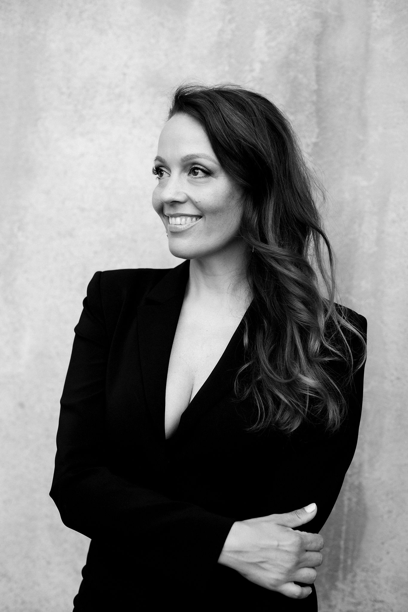 Black and white professional portrait of a businesswoman in a dark blazer with wavy hair smiling against a plain wall.