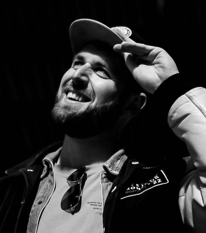 Black and white photo of a smiling bearded man wearing a cap and jacket, looking up while touching his hat.