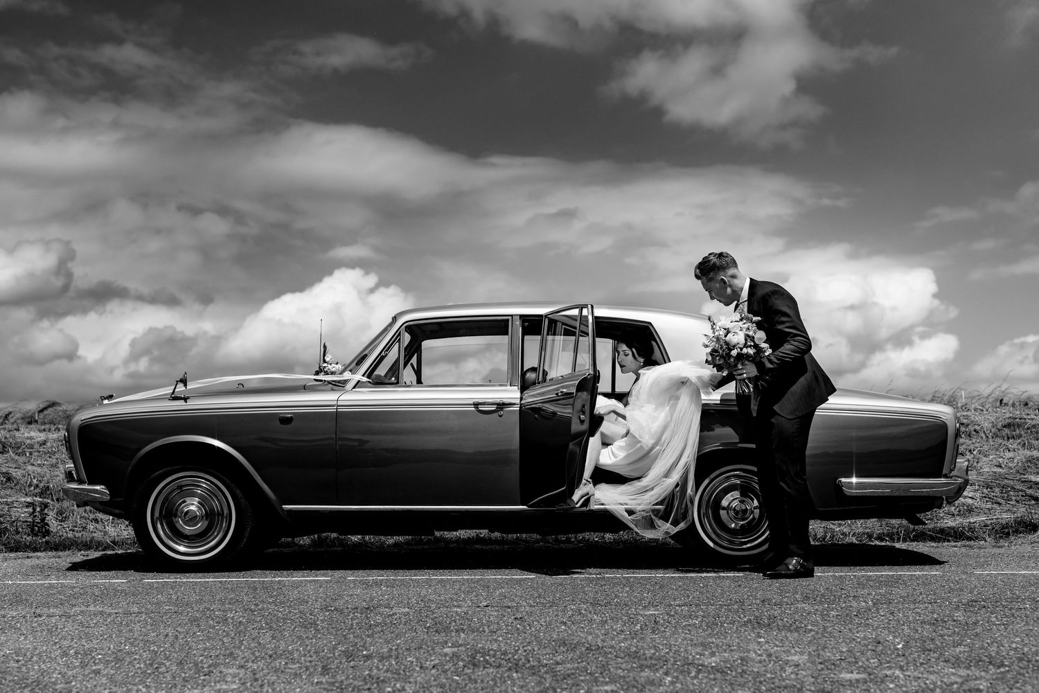 Classic vintage car on countryside road with two people in formal attire getting in, captured in black and white photography.