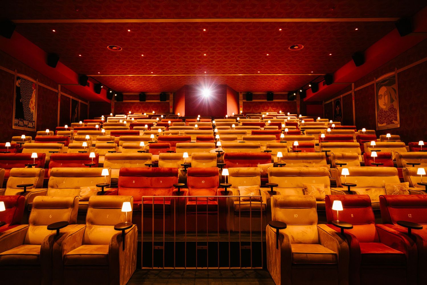Luxurious red cinema with recliner seats, small tables with warm lights, and a glowing red ceiling in a premium movie theater.