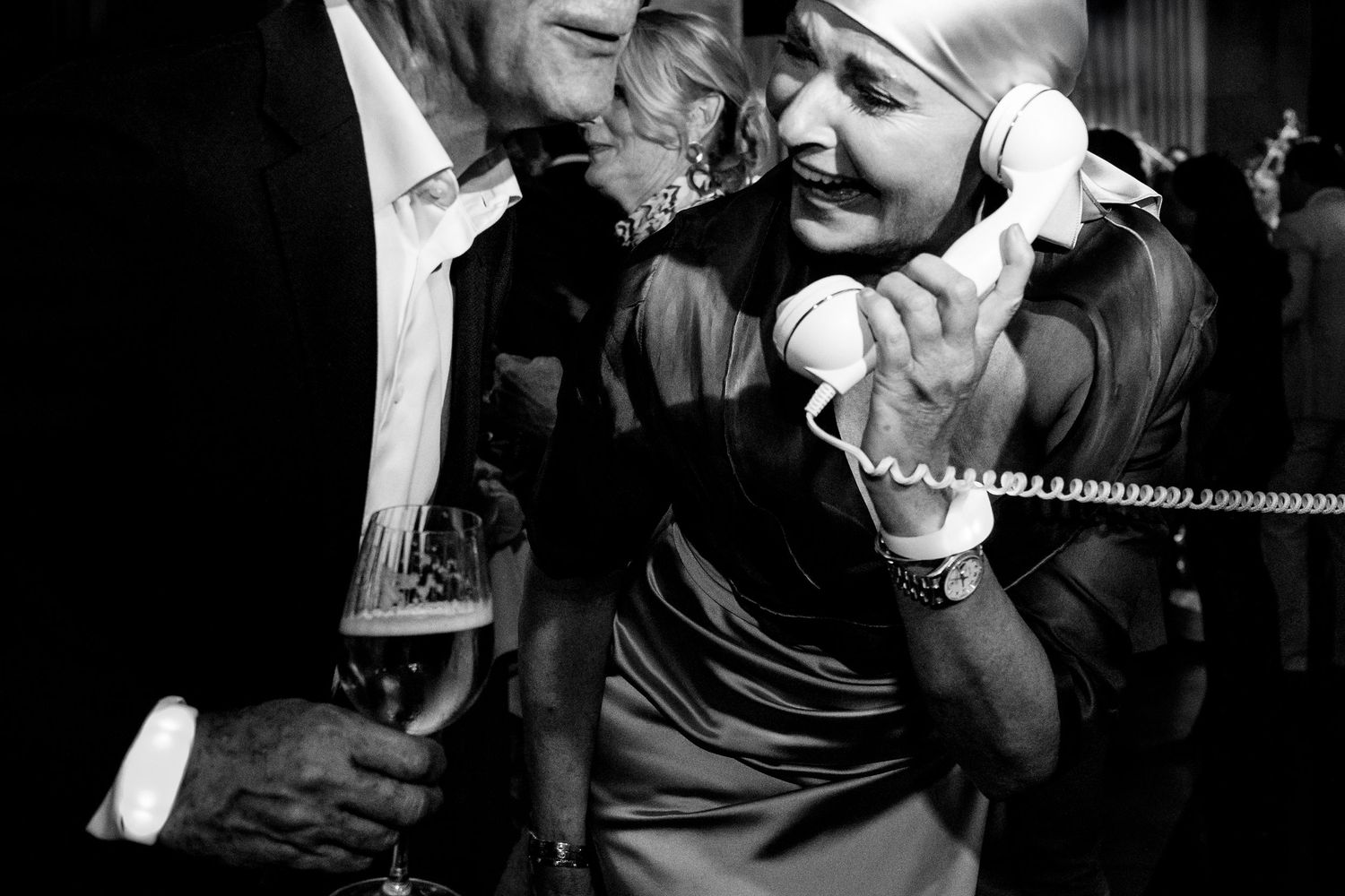 A black and white candid photograph of people laughing together while one holds a phone receiver at a party.