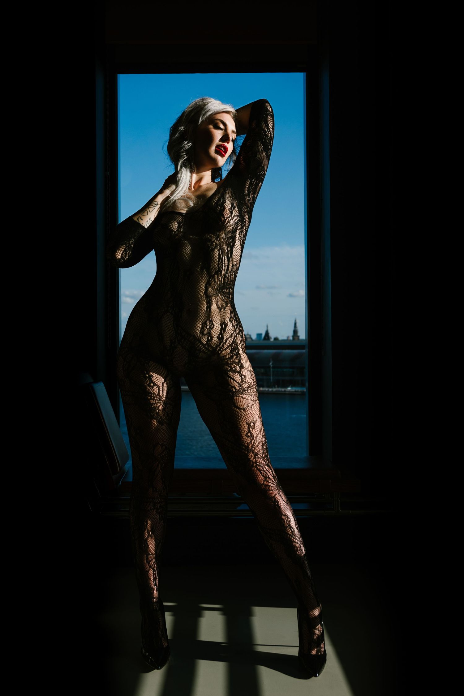 Silhouetted figure poses dramatically in lace bodysuit against window with blue sky background.