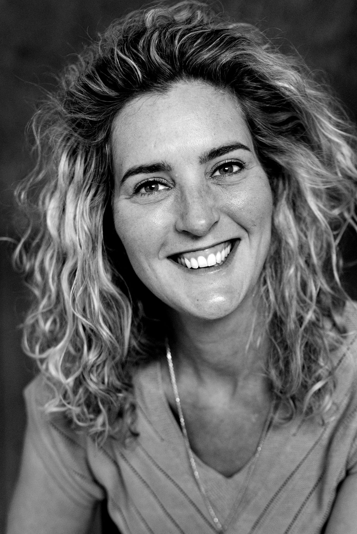 Black and white portrait of a smiling woman with curly blonde hair wearing a v-neck top and delicate necklace.