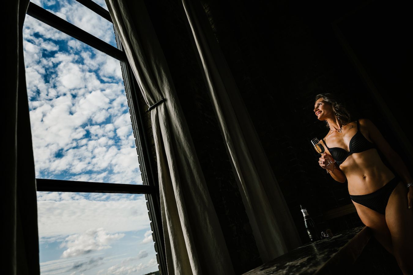 Woman in black lingerie holding champagne glass stands by large window with dramatic cloudy sky backdrop.