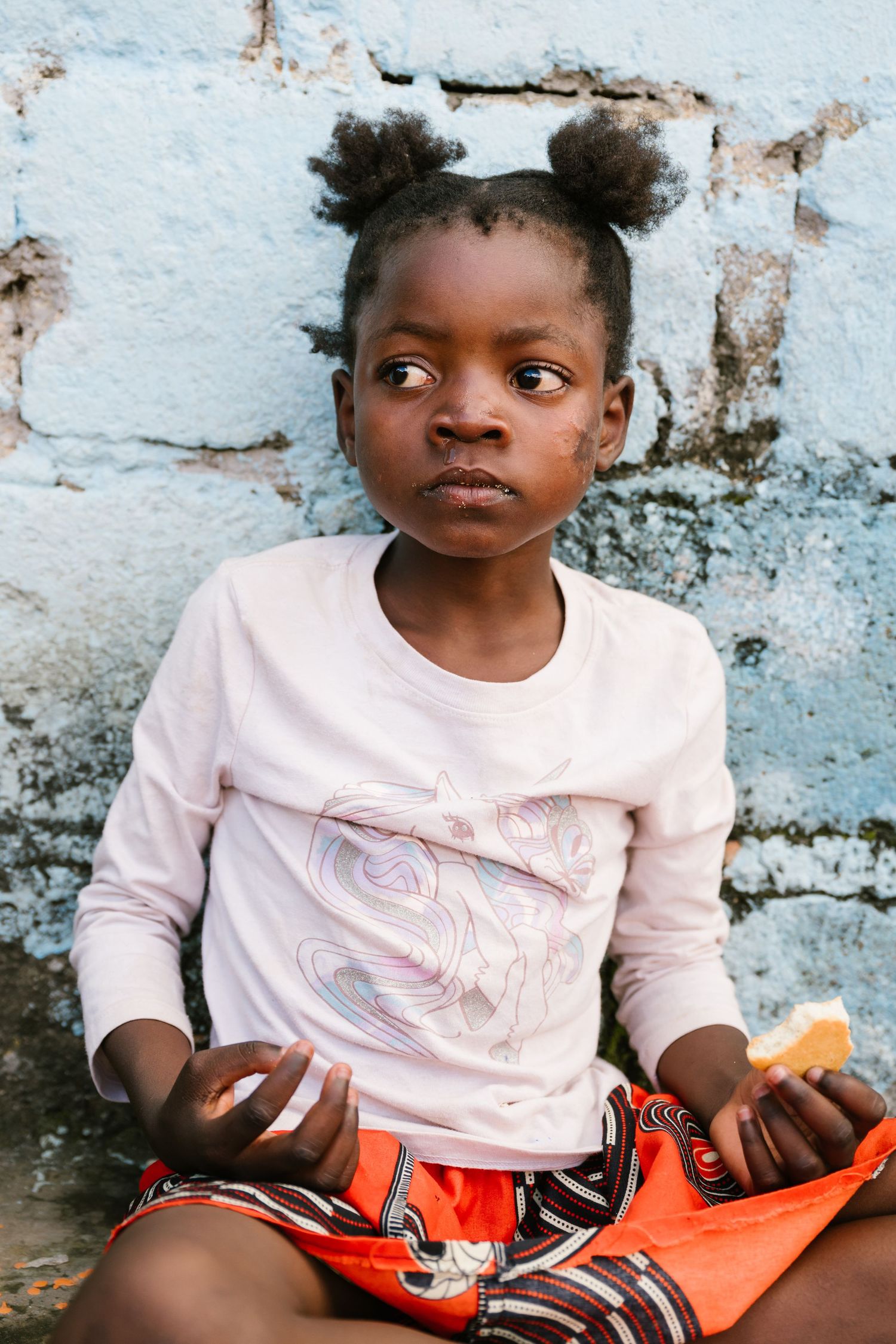 Young African girl with bun hairstyle wearing pink top and orange shorts, sitting against a weathered blue wall.