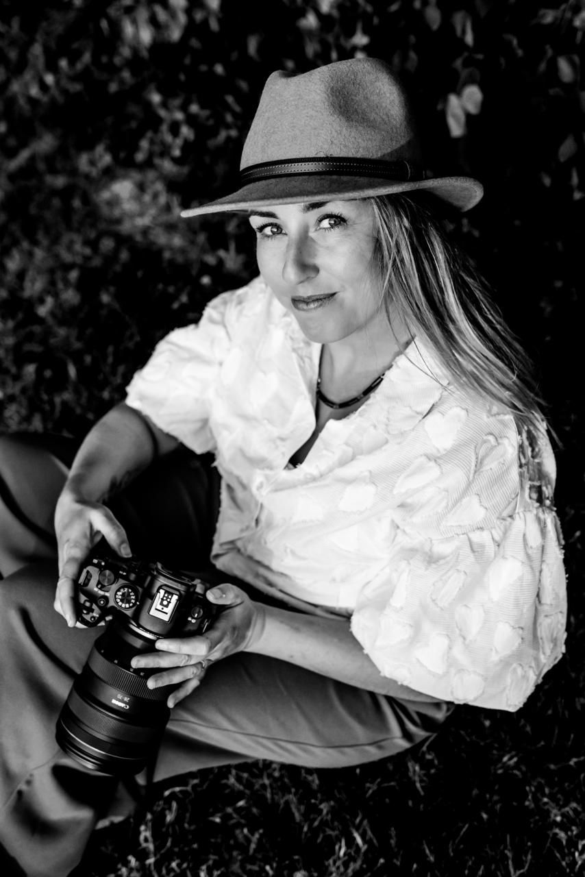 Black and white portrait of a blonde woman in a fedora hat holding a camera, sitting outdoors among green foliage.