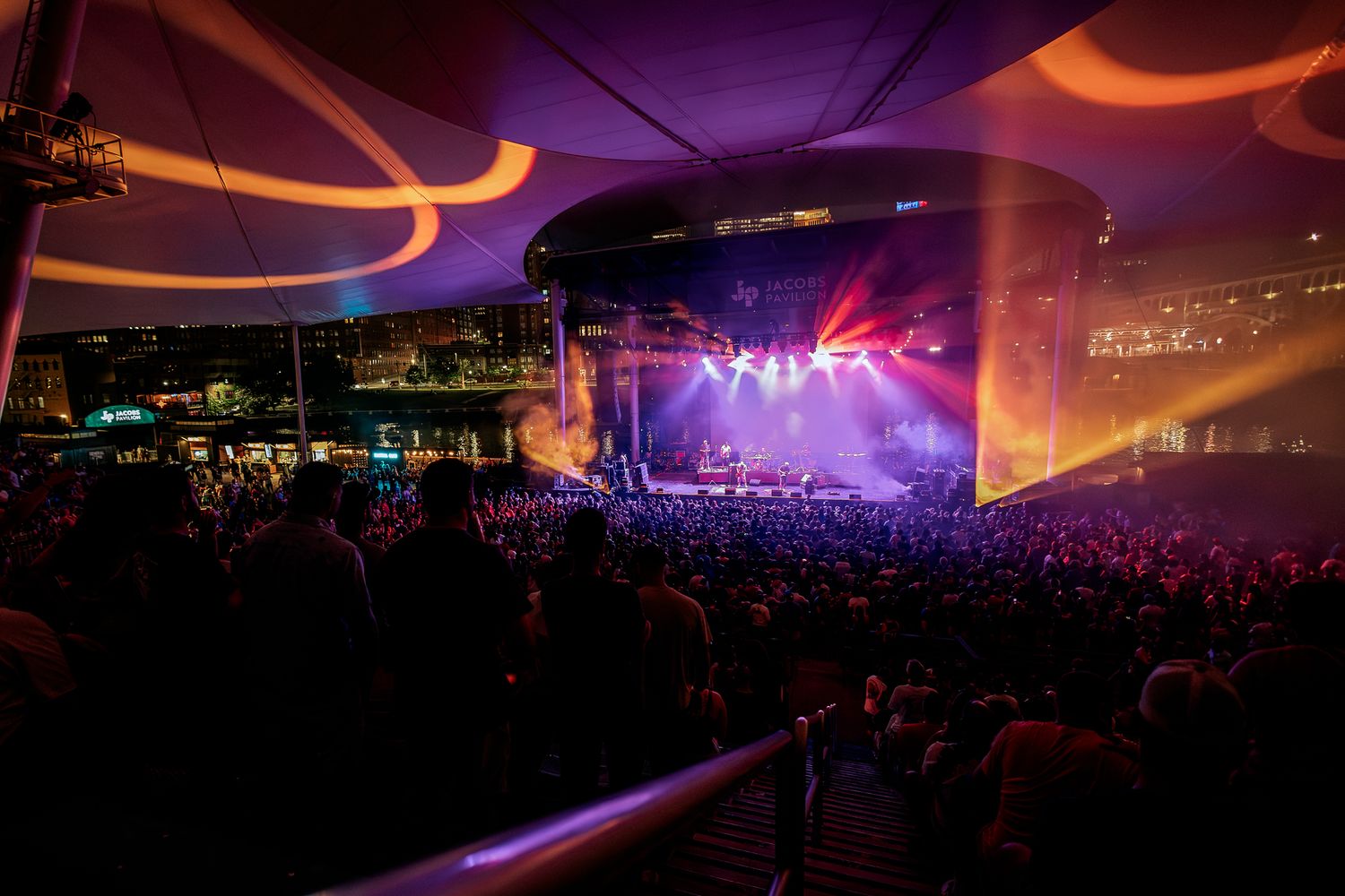 A concert venue with orange lighting and a packed crowd watching a performance on stage.