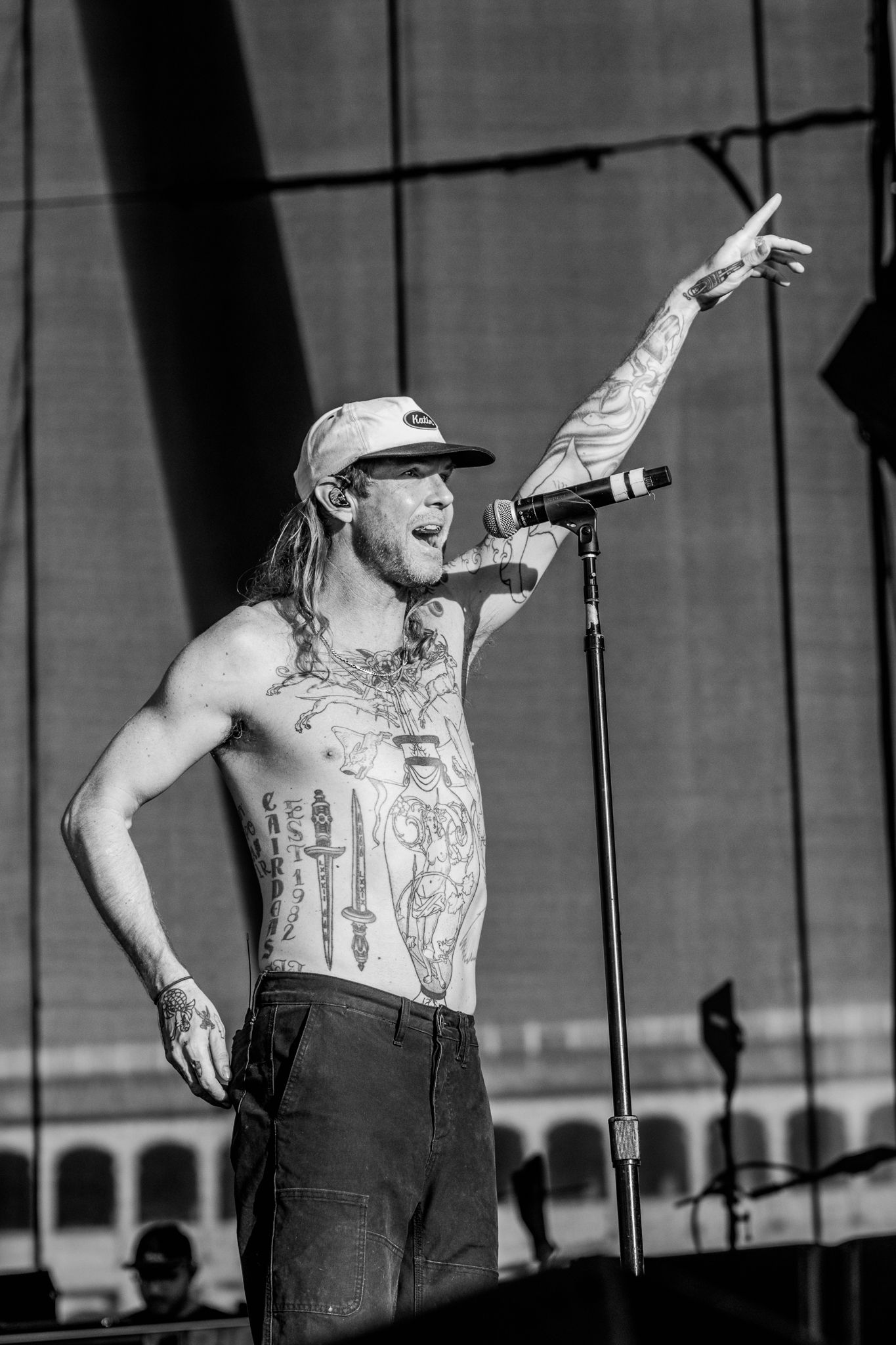 Shirtless performer gestures dramatically on outdoor concert stage in black and white.