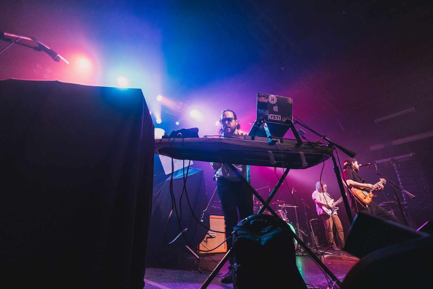 Concert stage setup with colorful pink and blue lighting showing electronic music equipment.