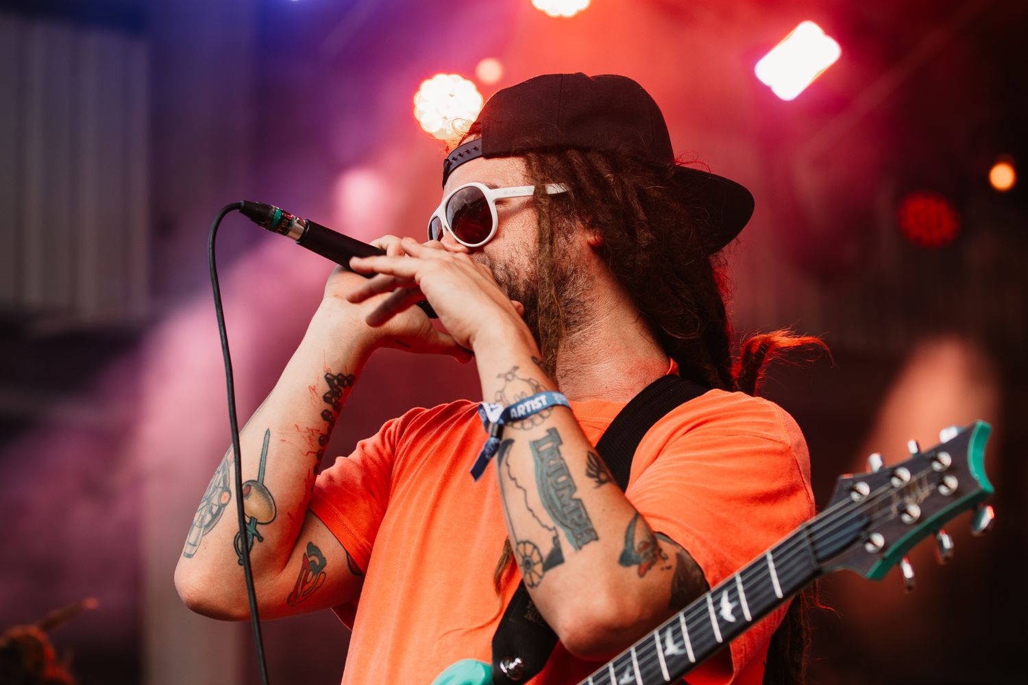 Musician in orange shirt performs with guitar against dramatic red stage lighting.