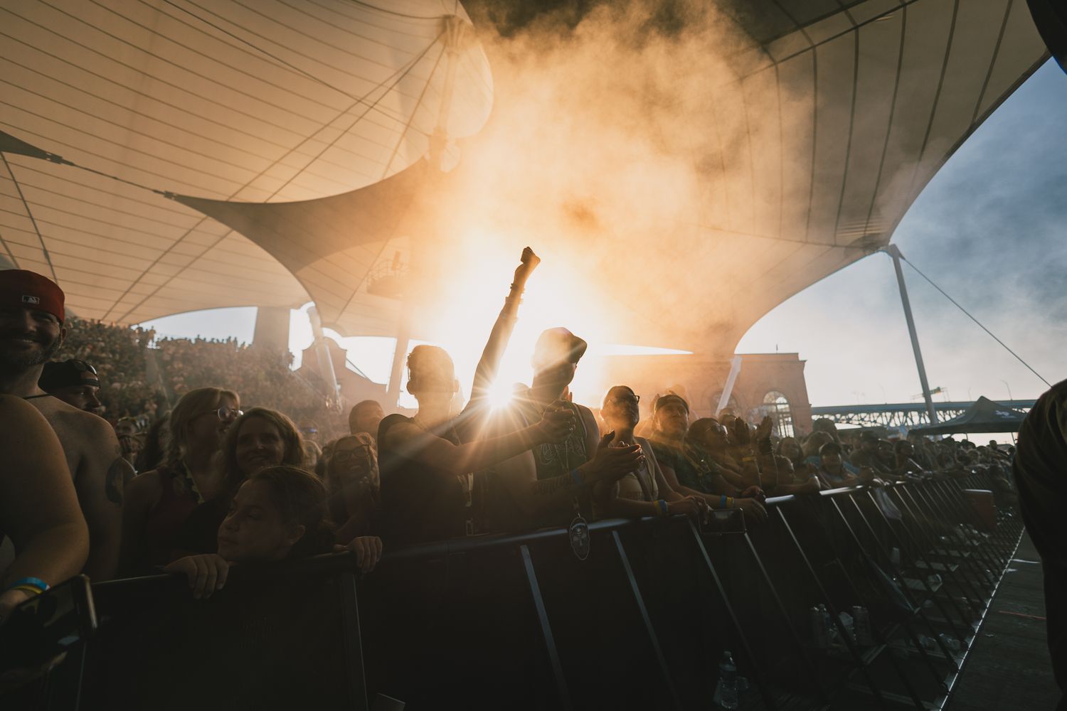 Silhouetted crowd at outdoor music festival raising hands against sunset-lit canopy.