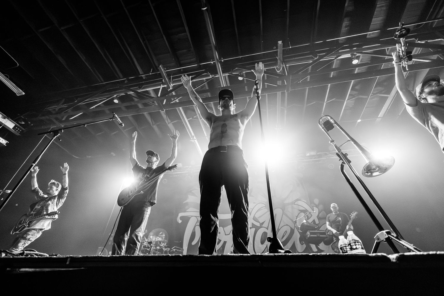 Silhouetted performers on stage raise their arms during a dramatic concert performance with bright backlighting.