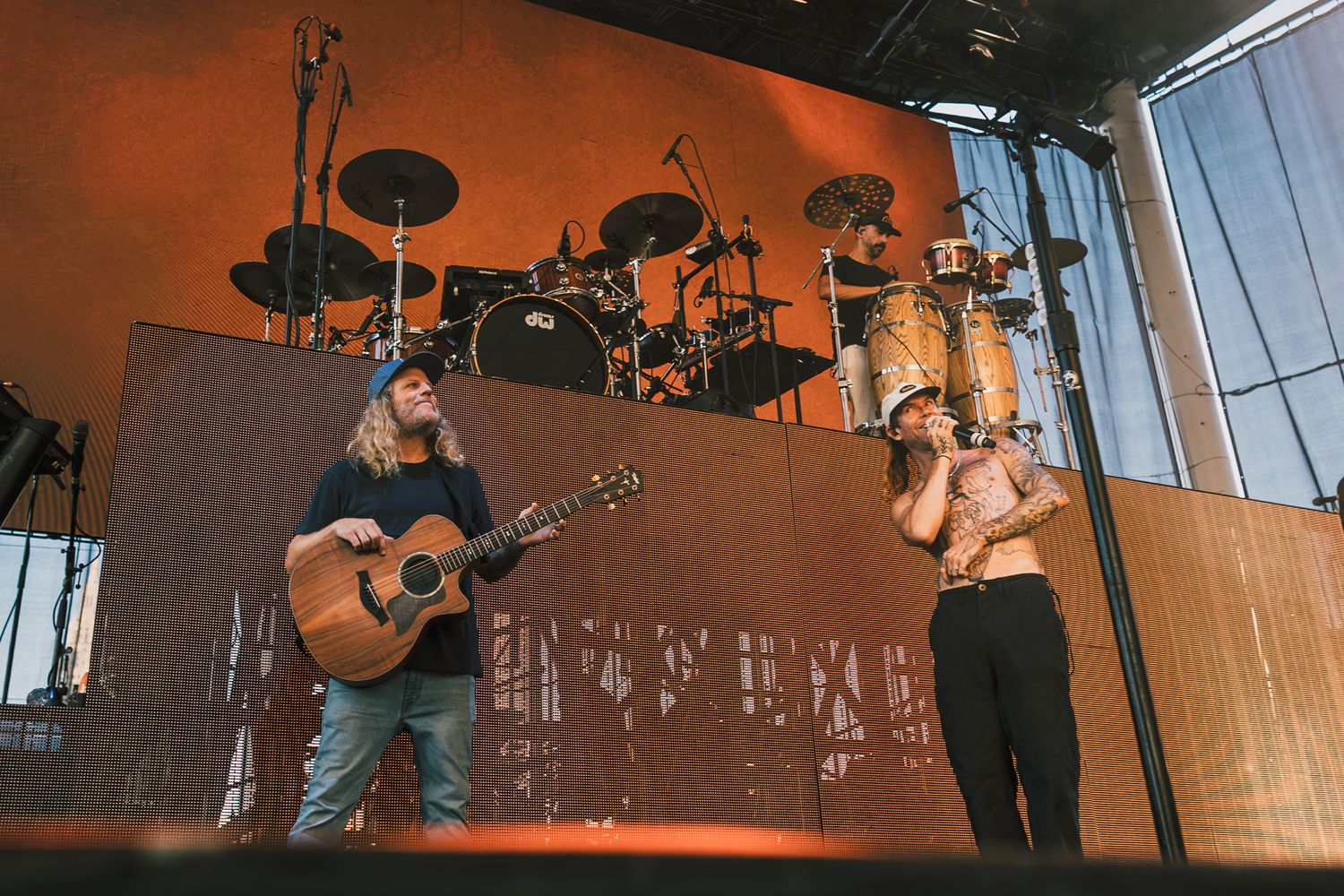 Musicians performing on outdoor stage with acoustic guitar and drum kit setup.