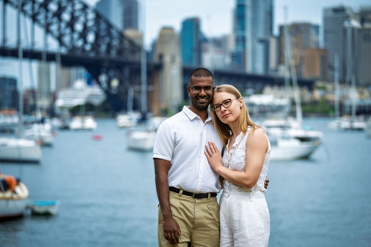 Couple poses affectionately near Sydney Harbour Bridge with boats in background.