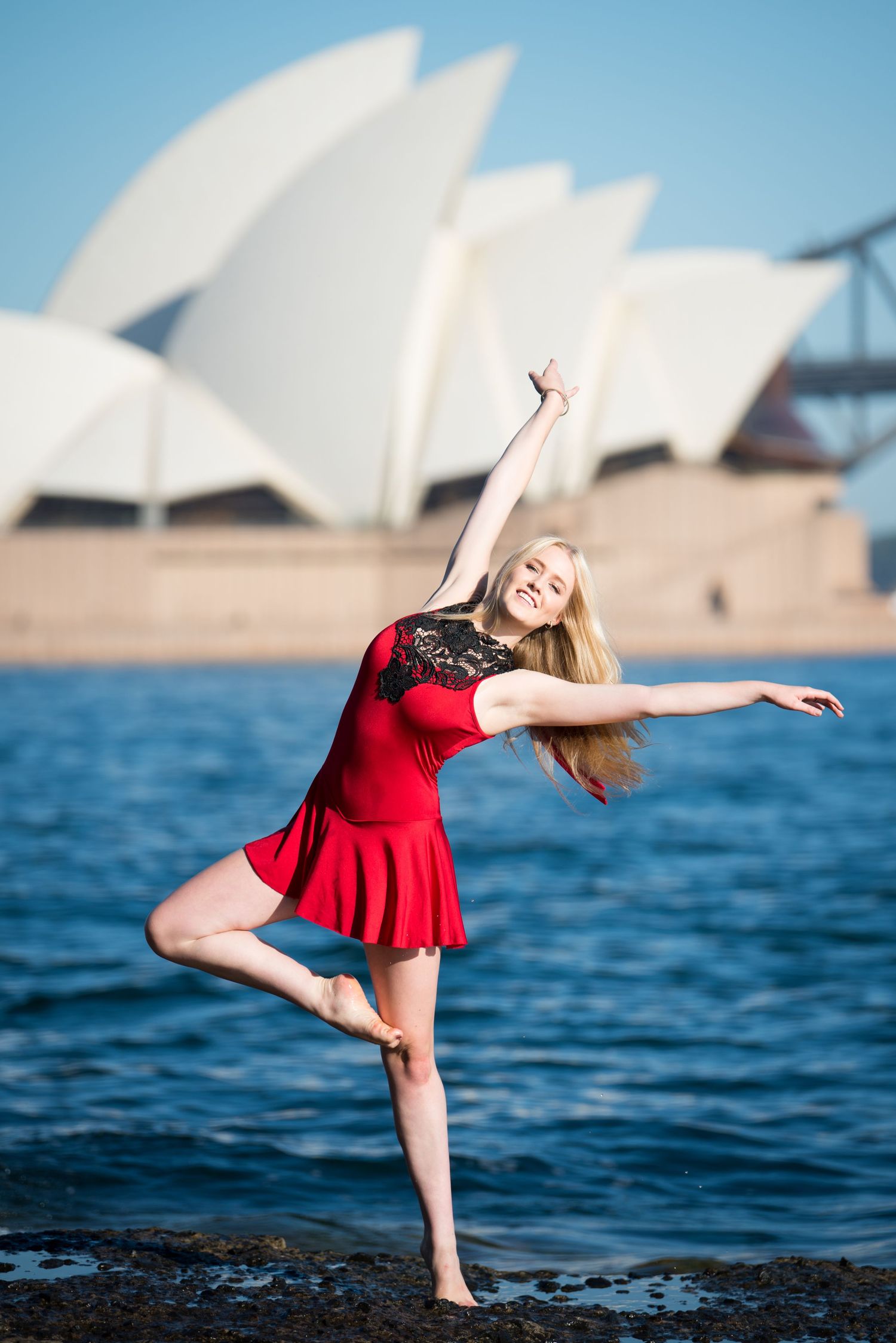 Dancer in red dress performs near Sydney Opera House with dramatic backdrop.