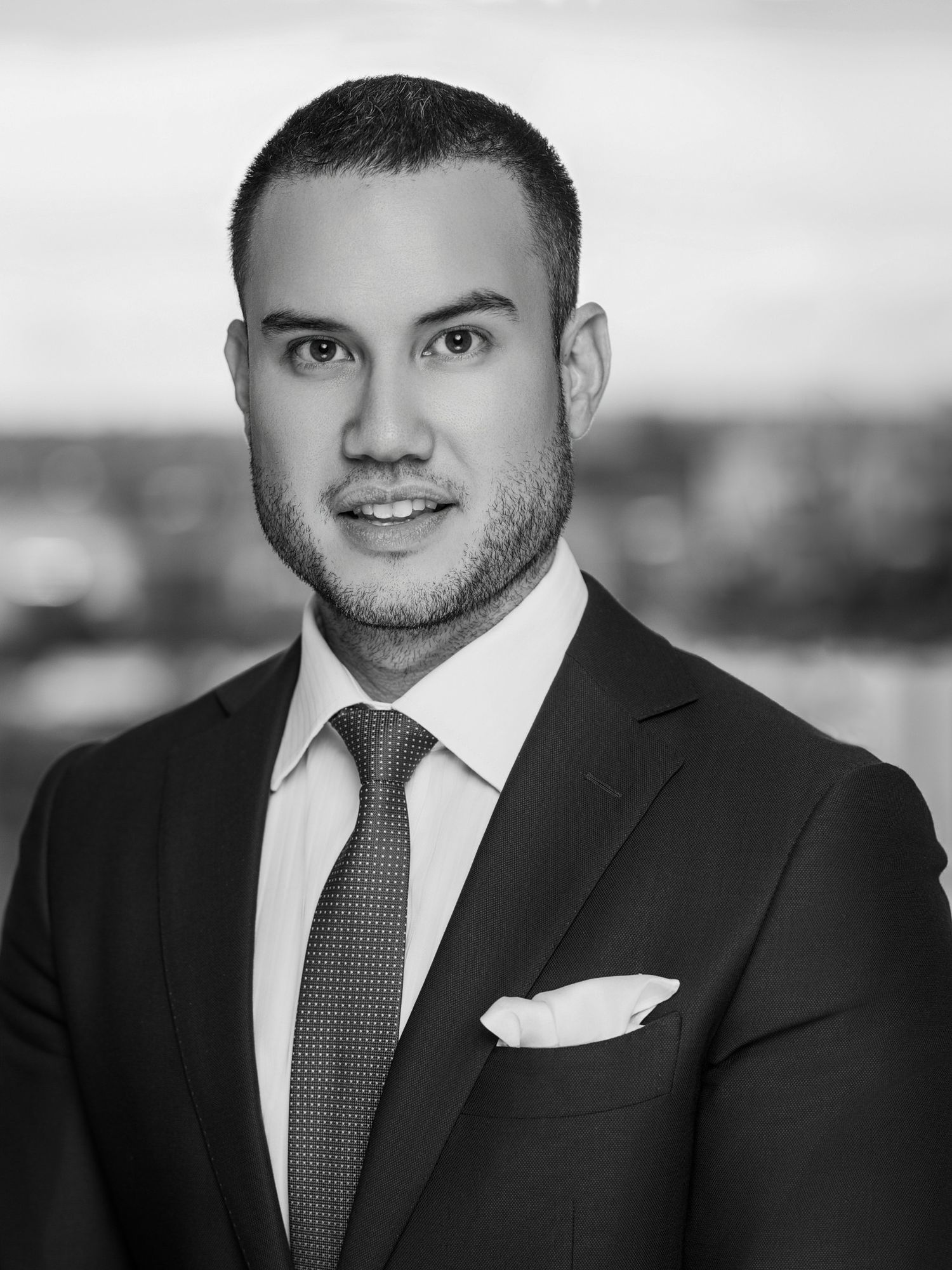 Professional headshot of person wearing dark suit with grey tie against light backdrop.